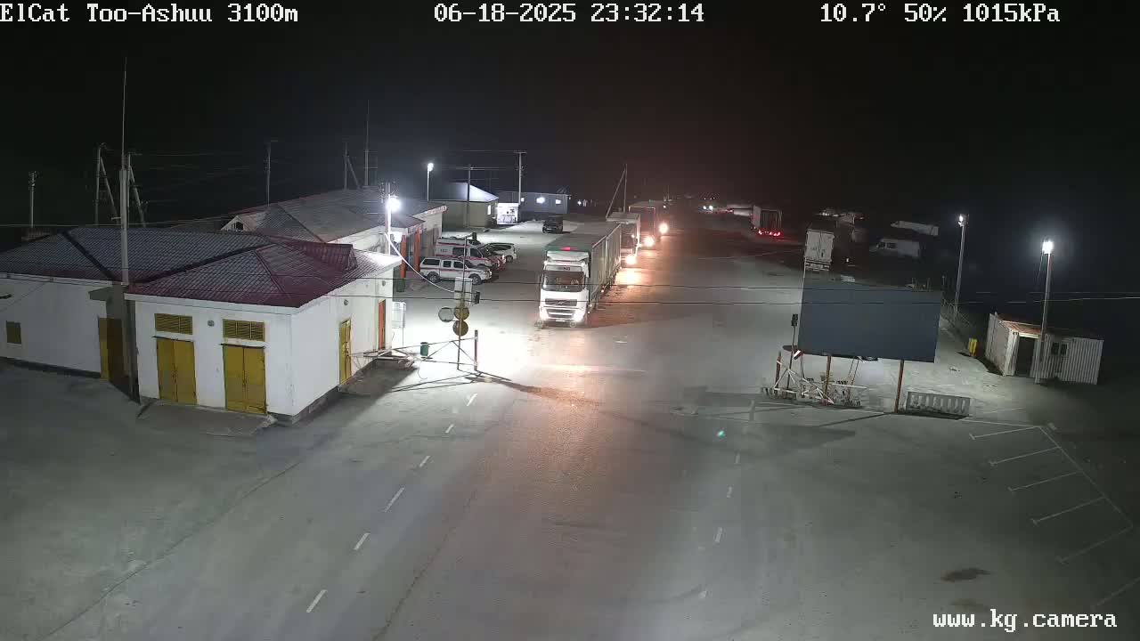 A nighttime, outdoor view of a roadway with several trucks and cars parked near a building under partly cloudy skies.