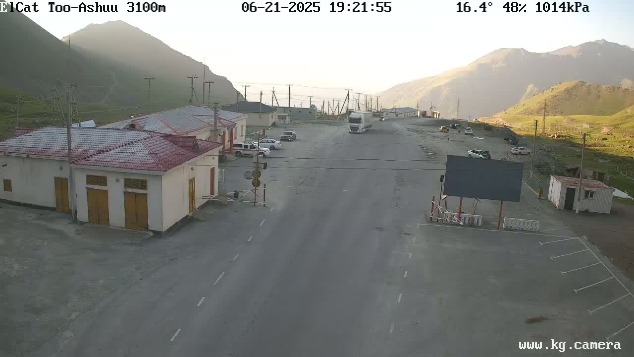 A mountain pass road scene shows a few cars and a truck at a roadside building complex under a partly sunny sky.