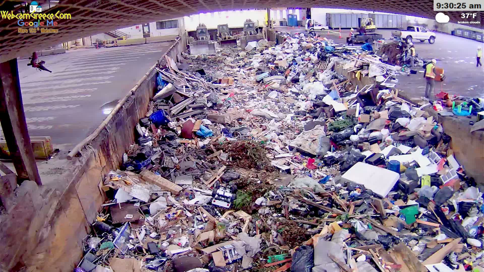 A large pile of trash and debris sits in an open-air waste receptacle under a partly cloudy sky, with several workers visible nearby and heavy machinery in the background.