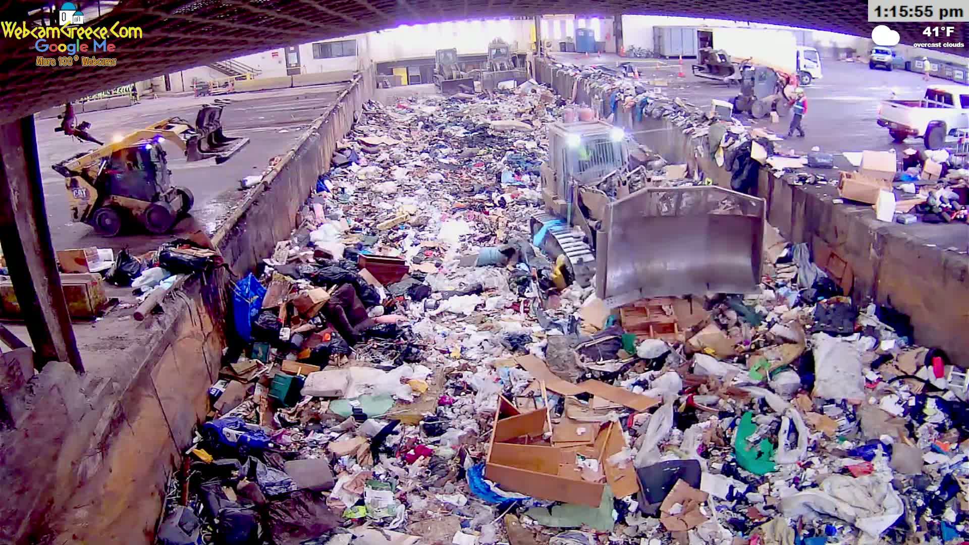 A large pile of garbage is being compacted by machinery in an outdoor area under an overcast sky.