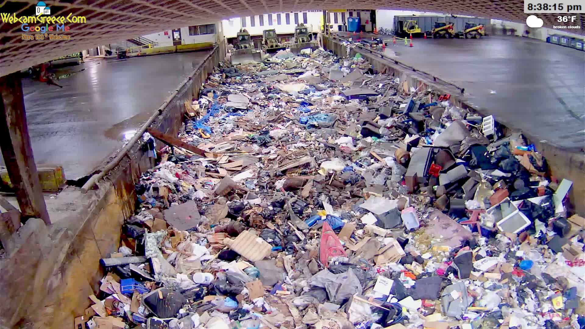 A large pile of trash sits in an open-air concrete container under a partially covered structure on a broken cloudy day with a temperature of 36°F.