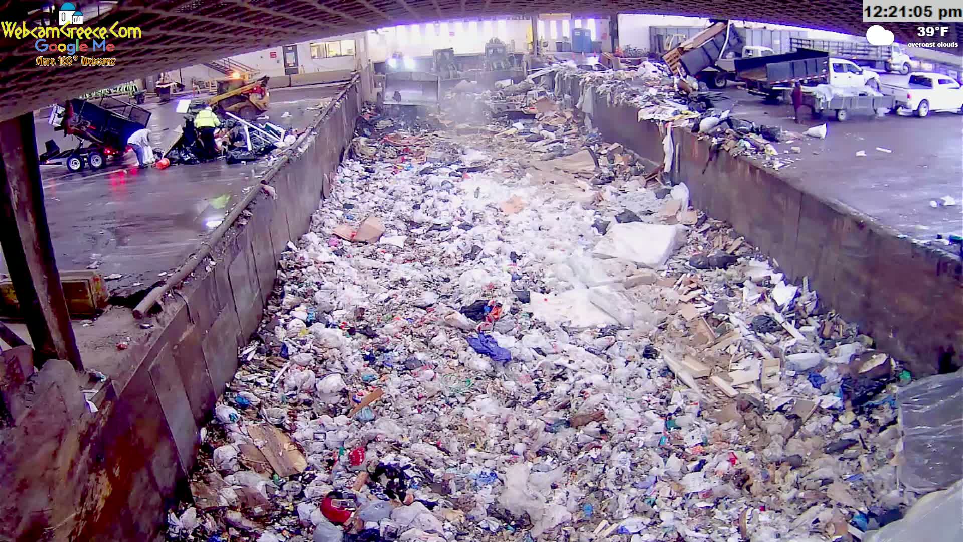 A large amount of garbage is piled in a trough-like structure outdoors under overcast skies, with some people and vehicles visible in the background.
