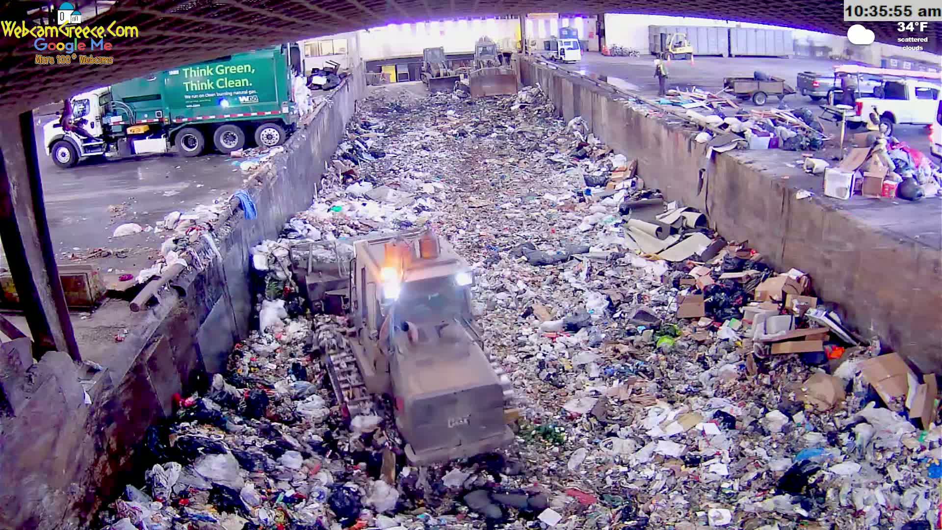 A bulldozer moves through a large pile of trash in an outdoor landfill under scattered clouds and a temperature of 34°F.