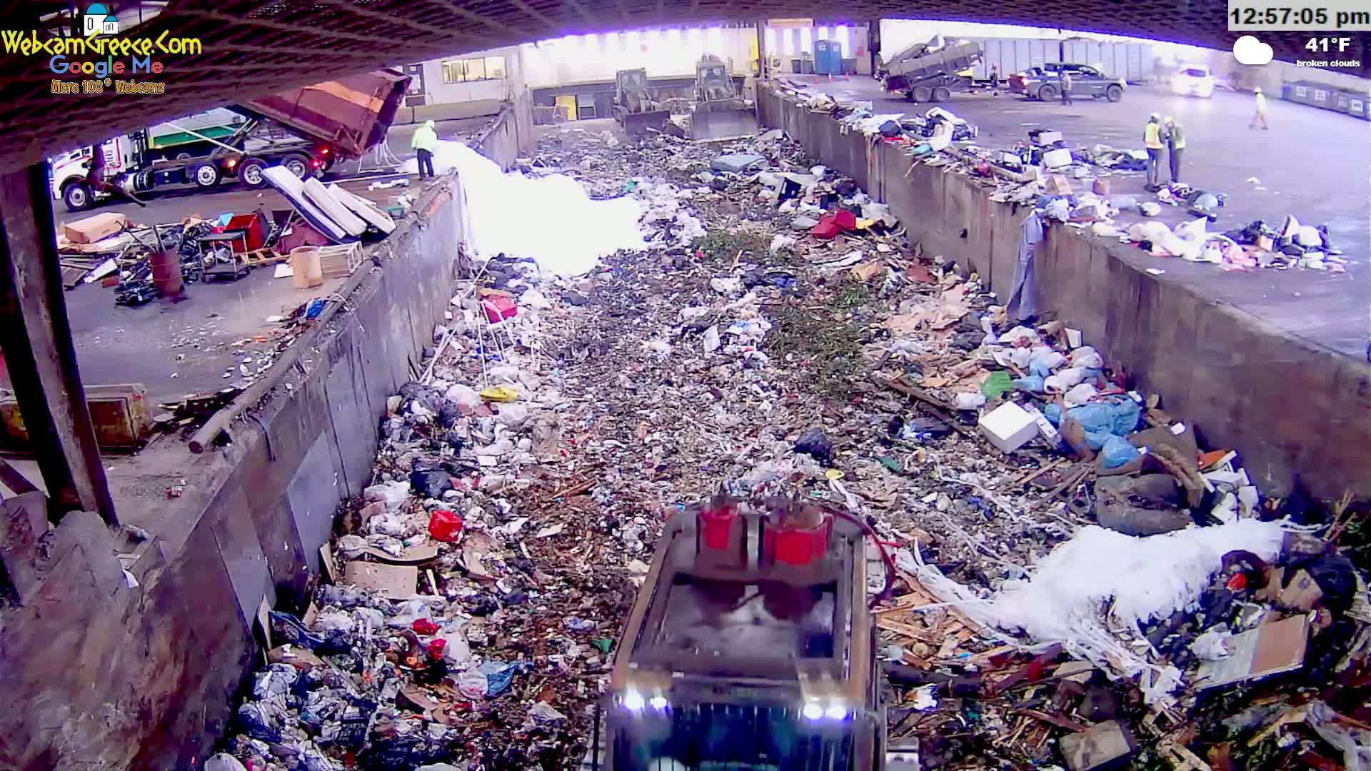 A large pile of trash and debris fills a waste disposal area under an overpass on a partly cloudy day.