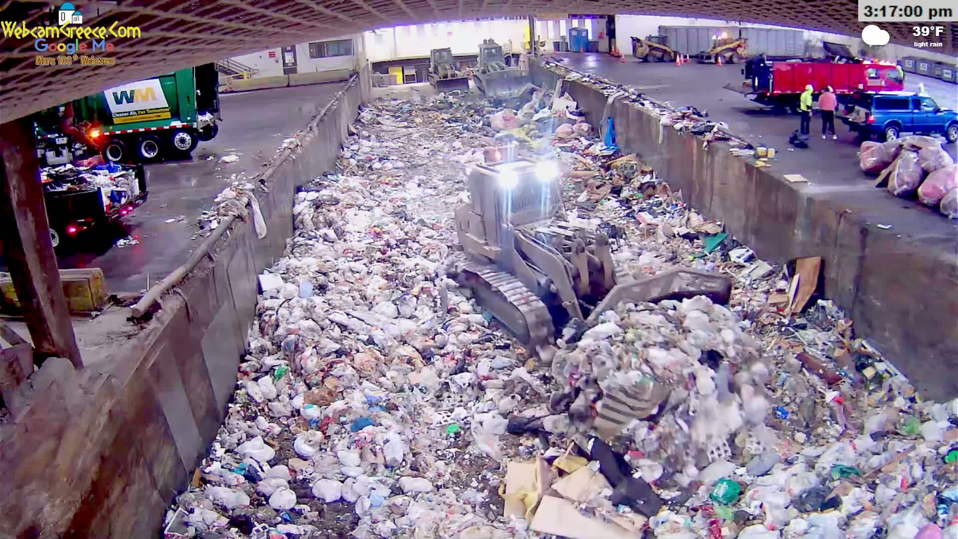 A small bulldozer pushes trash in a large, partially covered landfill during light rain.