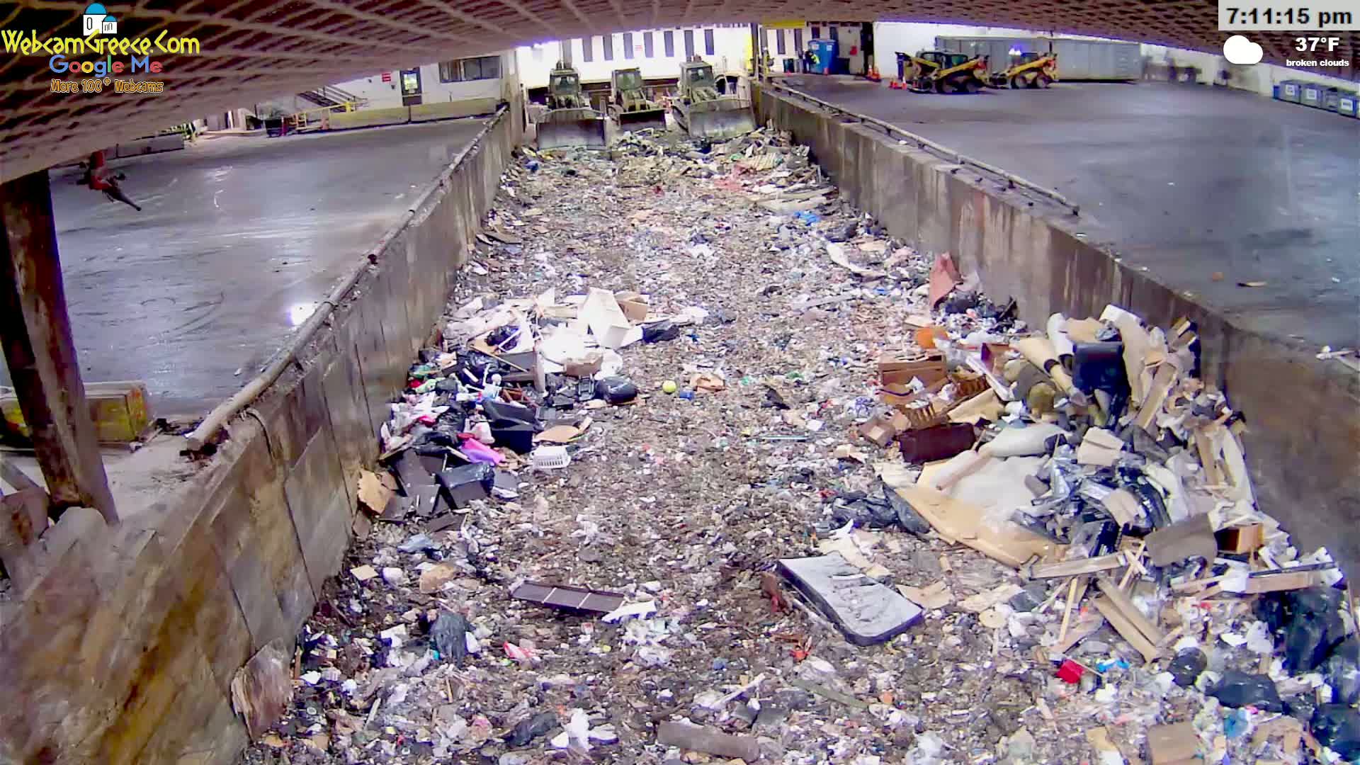 A large pile of trash sits in a concrete trough under a covered area, with two pieces of heavy machinery visible in the background under broken clouds and a temperature of 37°F.