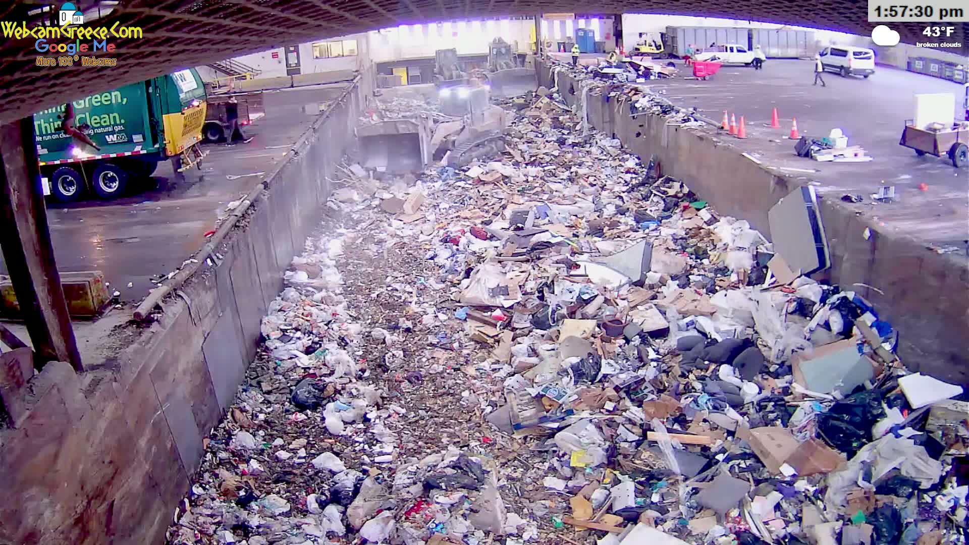 A large pile of trash sits in an outdoor, covered waste disposal area under broken clouds on a 43°F day, with a garbage truck and a bulldozer visible nearby.