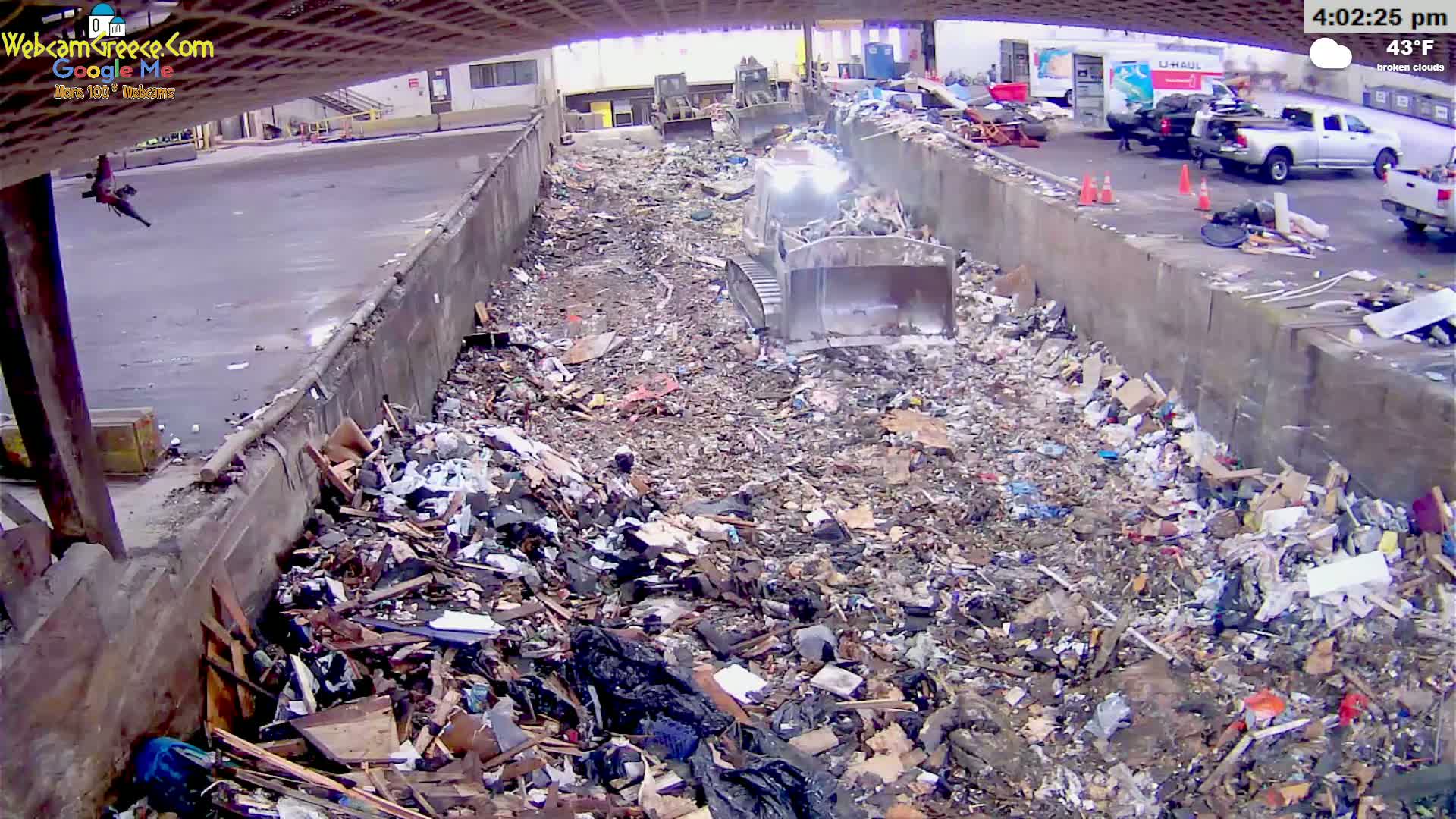 A large pile of trash is being compacted by machinery in a waste processing facility under broken clouds at 43°F.