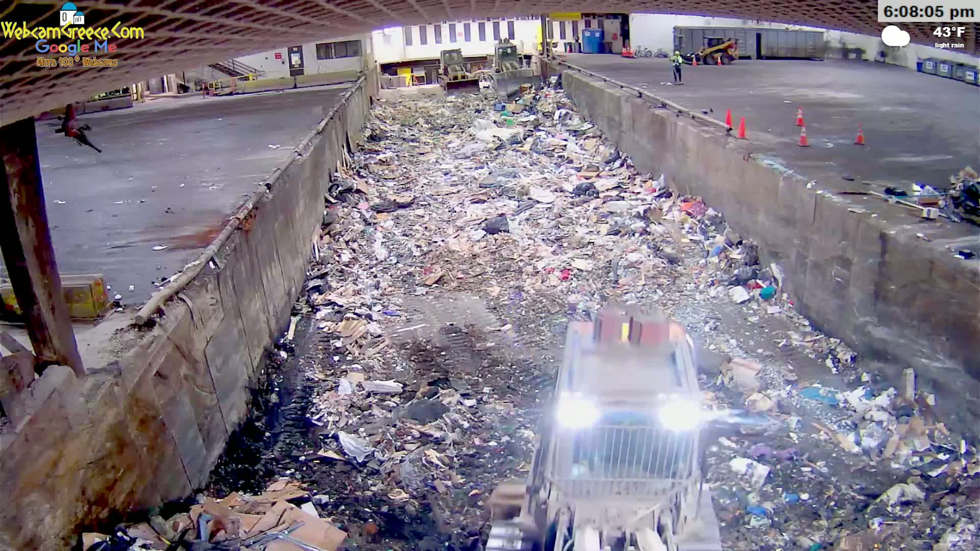 A large pile of trash is seen from an elevated view in a large, concrete-walled enclosure, with a garbage truck operating at the bottom of the pile under light rain.