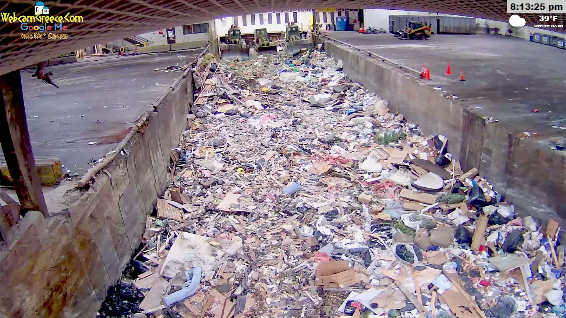 A large pile of trash sits in a concrete trough outdoors under overcast skies, with three pieces of heavy machinery visible in the background.