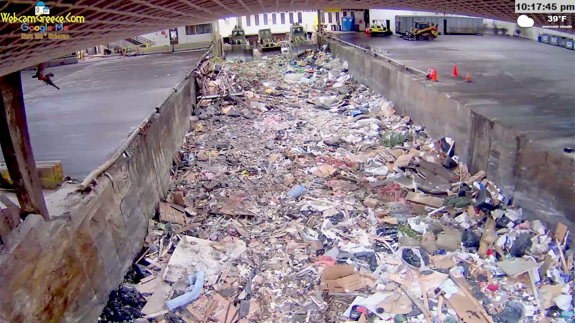 A large amount of refuse is piled in an outdoor concrete trough under overcast skies, with several pieces of heavy machinery visible in the background.