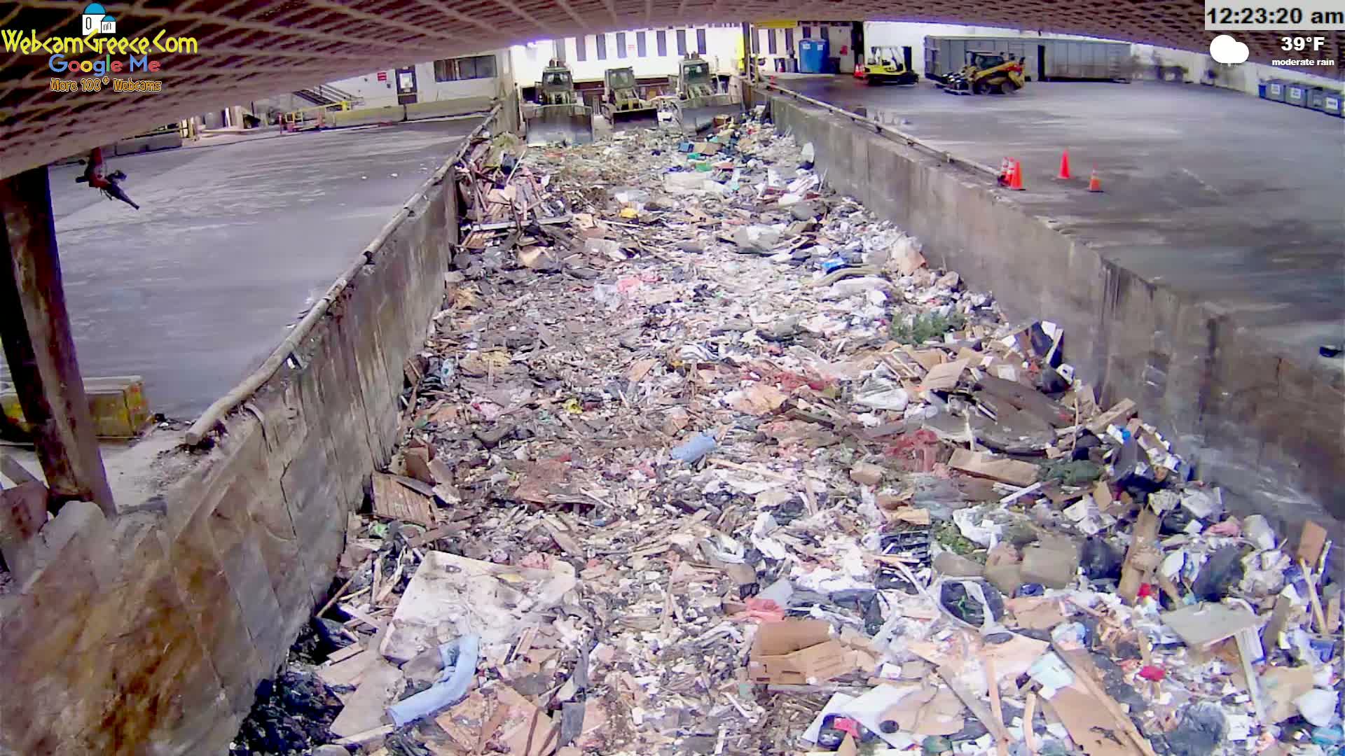 A large pile of trash fills a concrete trough under a partly covered structure, with three pieces of heavy machinery visible in the background on a moderately rainy 39°F day.