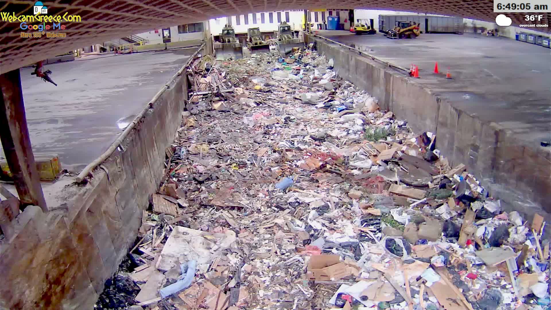 A large pile of trash fills a concrete trough under an overcast sky, with three pieces of heavy machinery visible in the background.