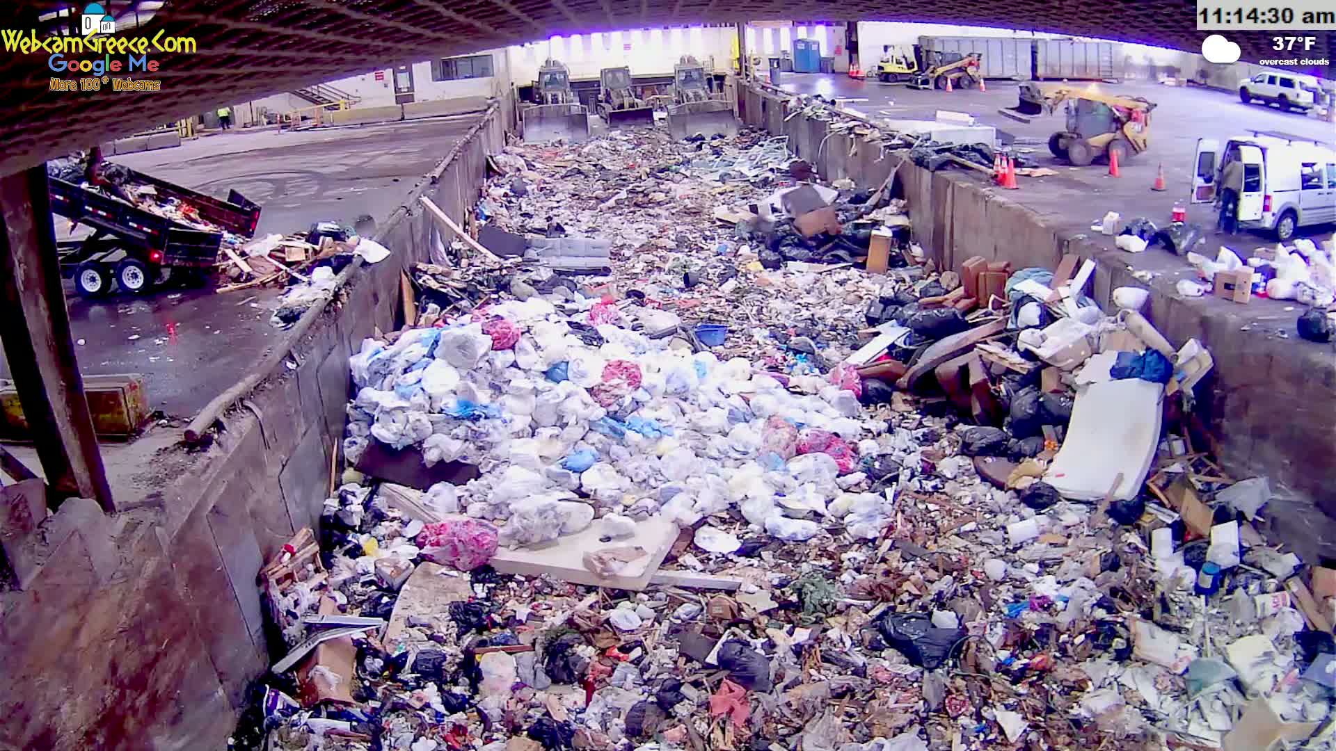A large pile of trash sits inside a waste receptacle under overcast skies, with a dump truck and other vehicles visible in the background.