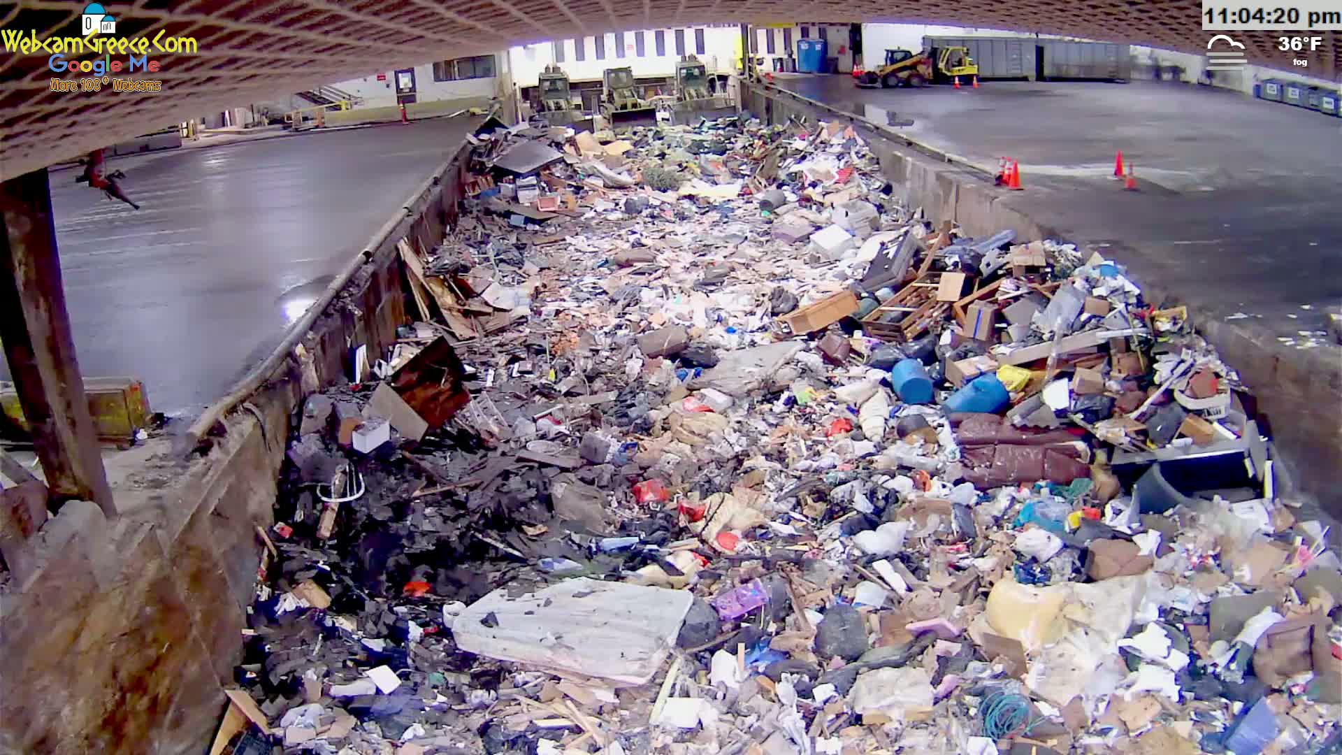 A large pile of refuse sits in an open-air, concrete waste receptacle under a foggy, 36°F sky.