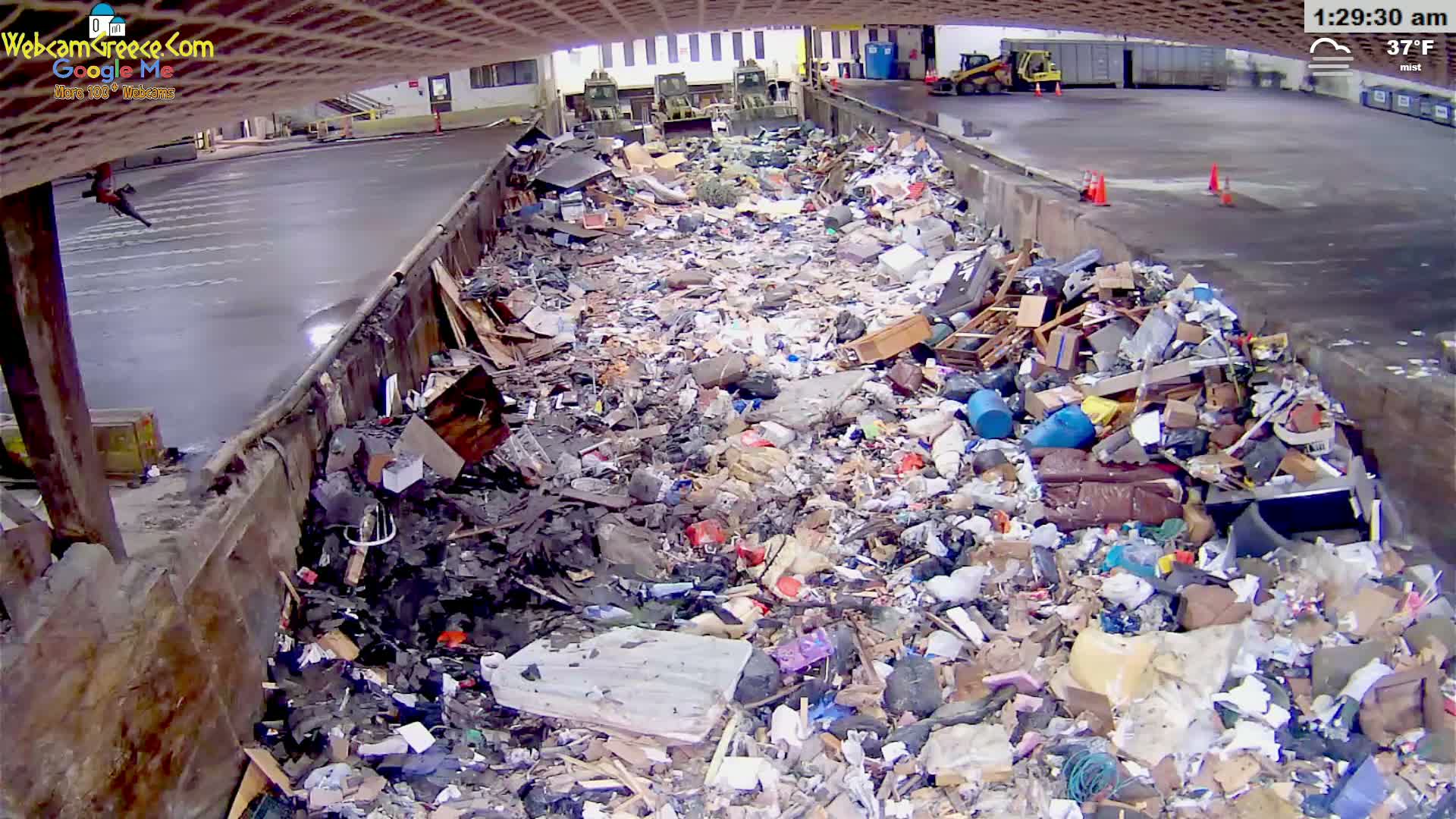 A large pile of refuse sits in a misty, outdoor, industrial-looking area with a paved surface.