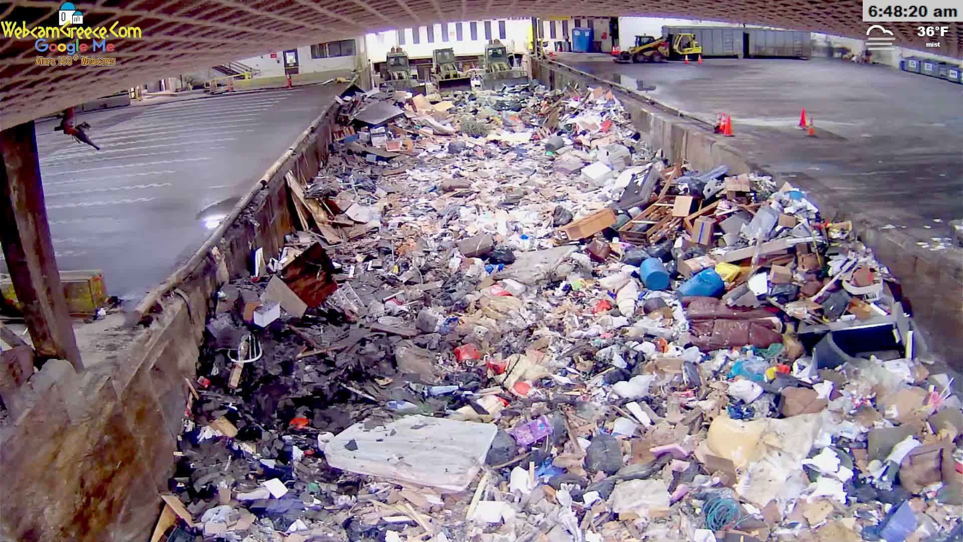 A large pile of trash and debris sits in an outdoor, mist-covered area next to a paved lot.
