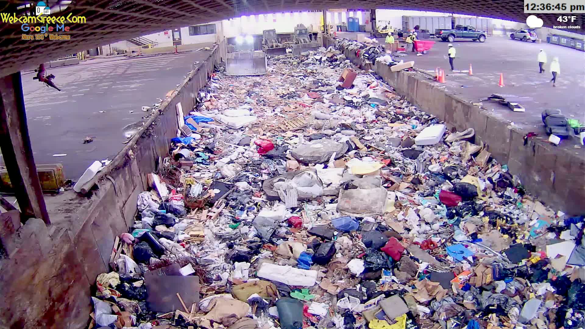 A large amount of garbage fills a long, open container in an outdoor area under a partly cloudy sky, with workers visible in the background.