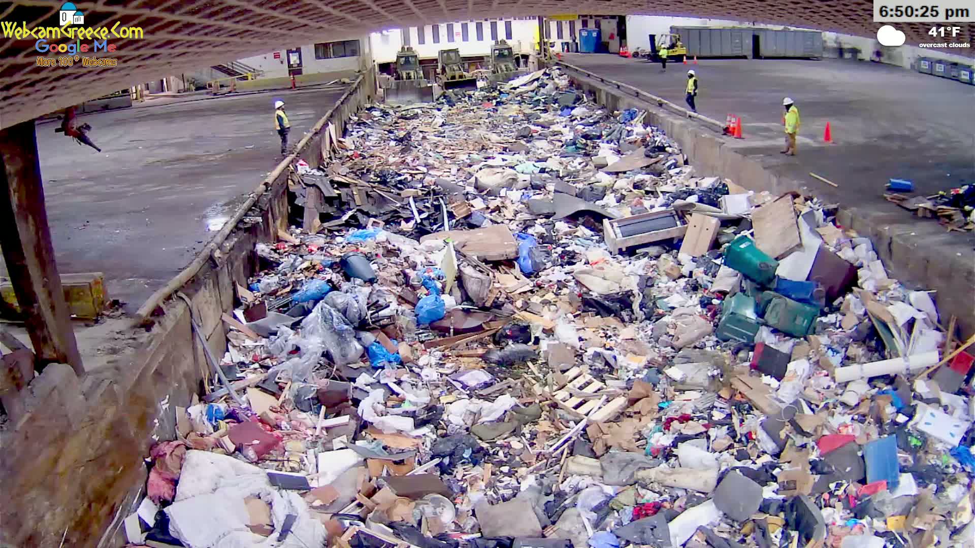 A large amount of refuse is piled high in an outdoor waste receptacle under overcast skies, with several workers visible nearby.