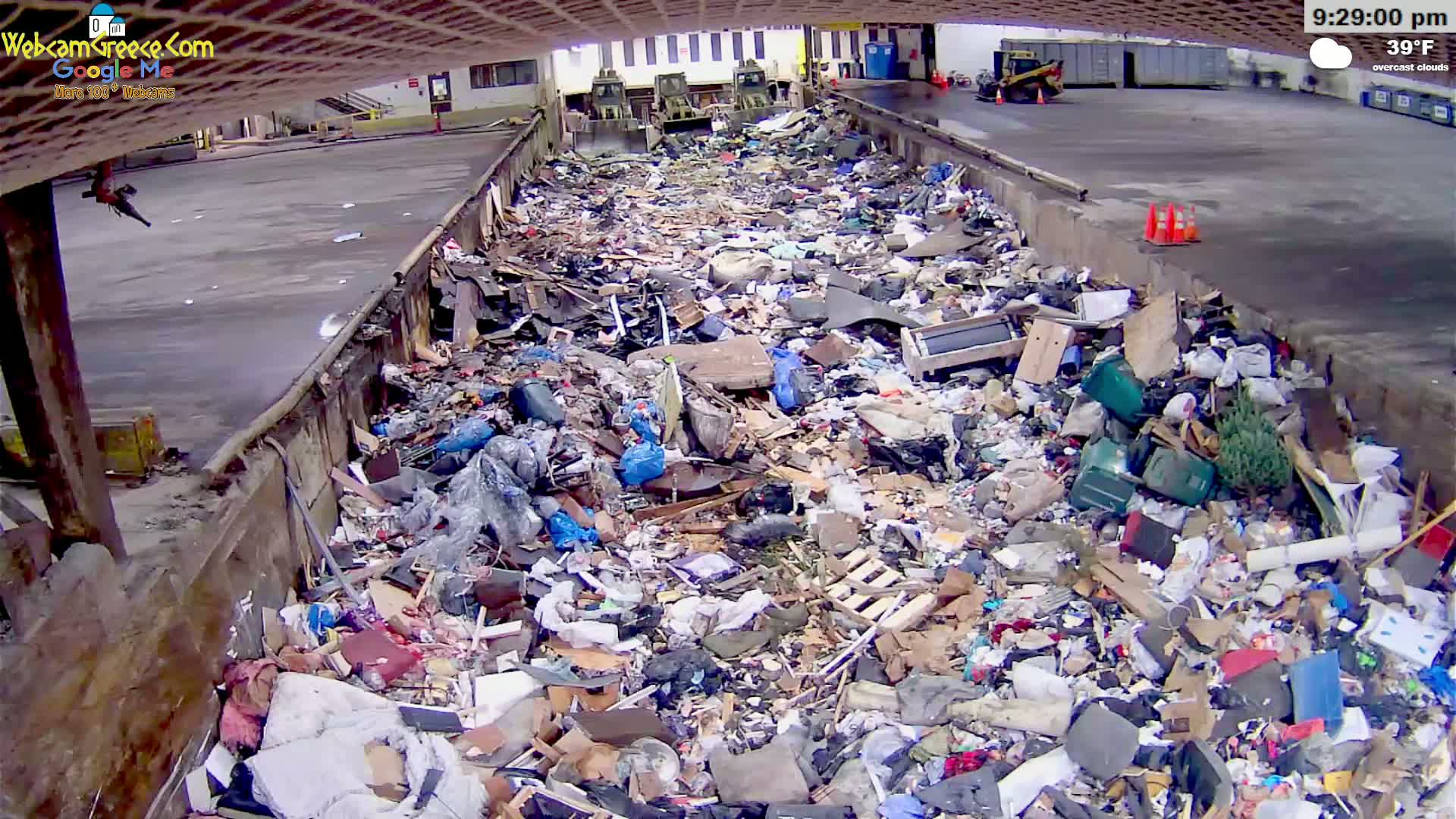 A large pile of trash sits in an outdoor, covered, concrete waste receptacle under overcast skies, with the temperature at 39°F.