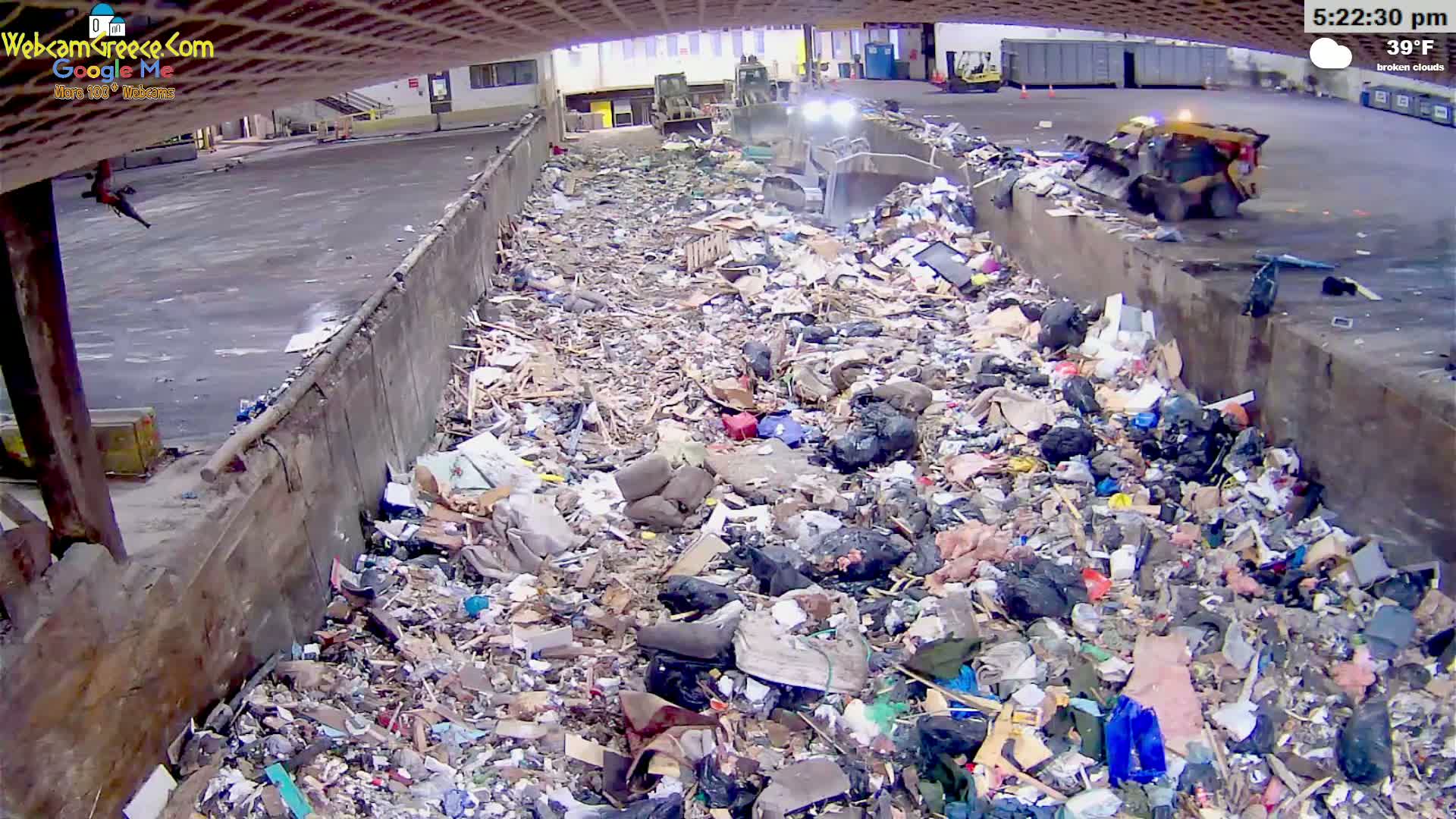 A large pile of trash is seen in an outdoor, open-air facility under broken clouds with a temperature of 39°F, with heavy machinery nearby.
