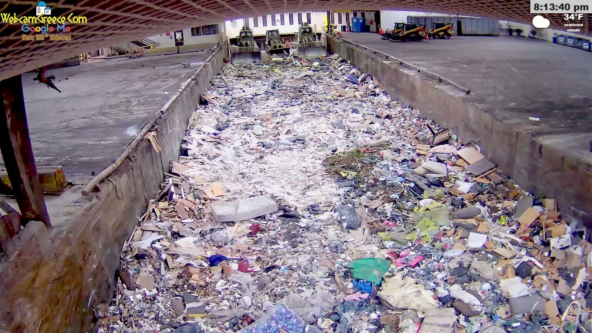 A large pile of trash fills a concrete-walled pit outdoors under scattered clouds and a 34°F temperature.