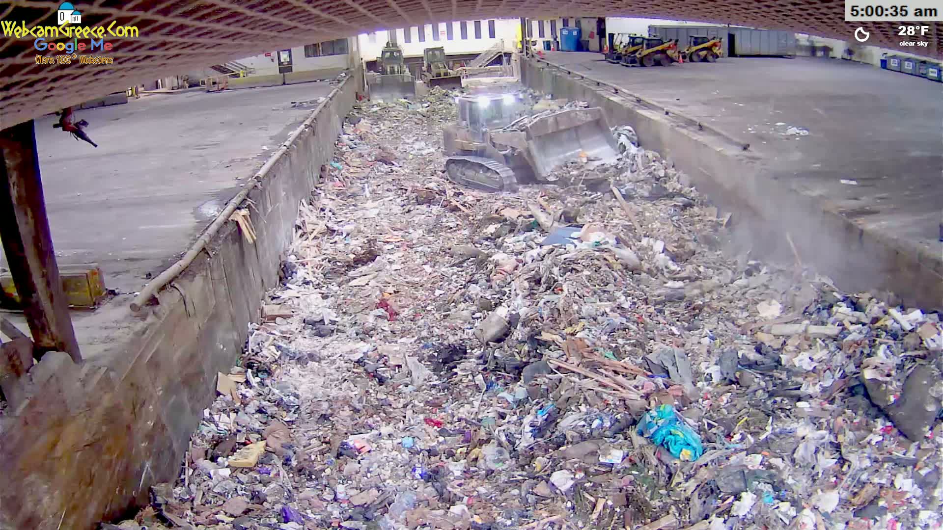 A bulldozer is compacting a large pile of trash in an outdoor waste disposal area under clear skies, with a temperature of 28°F.