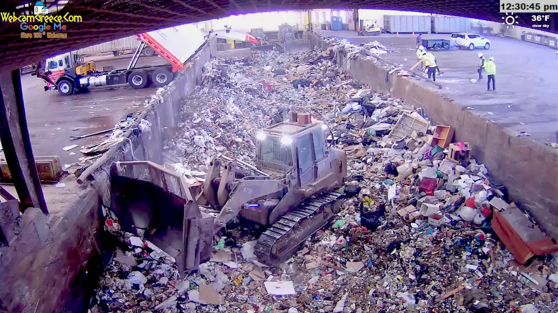 A bulldozer is working in a large pile of trash under a clear sky, with several people and a dump truck visible nearby.