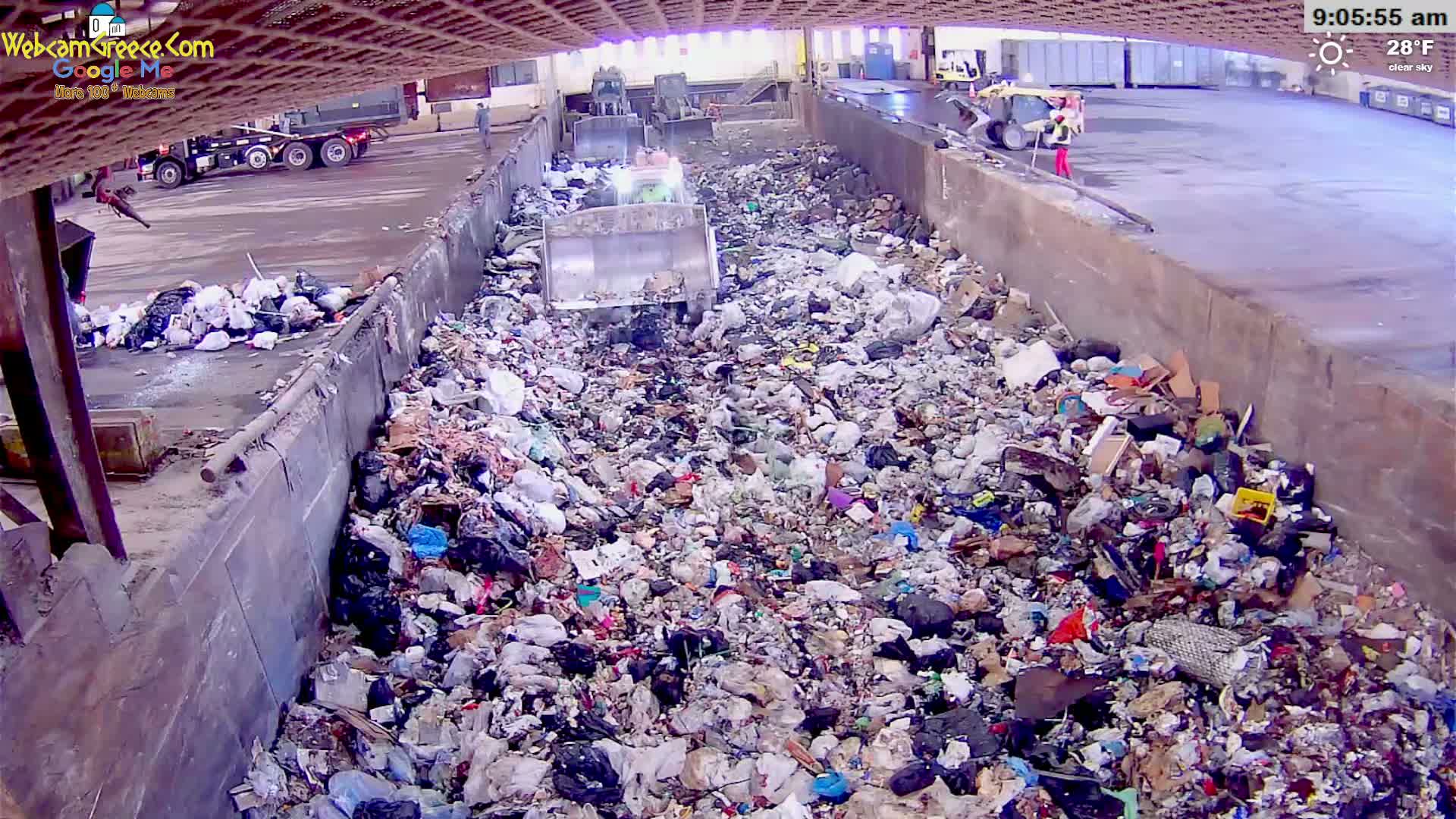 A large pile of garbage is being compacted by machinery in an outdoor facility under a clear sky on a 28°F day.
