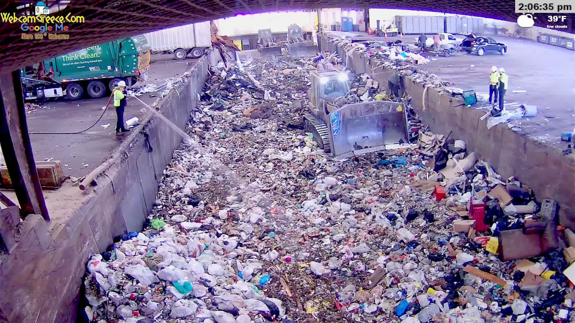 A large pile of trash is being compacted by a bulldozer in a waste management facility under a partially covered structure on a day with few clouds and a temperature of 39°F.