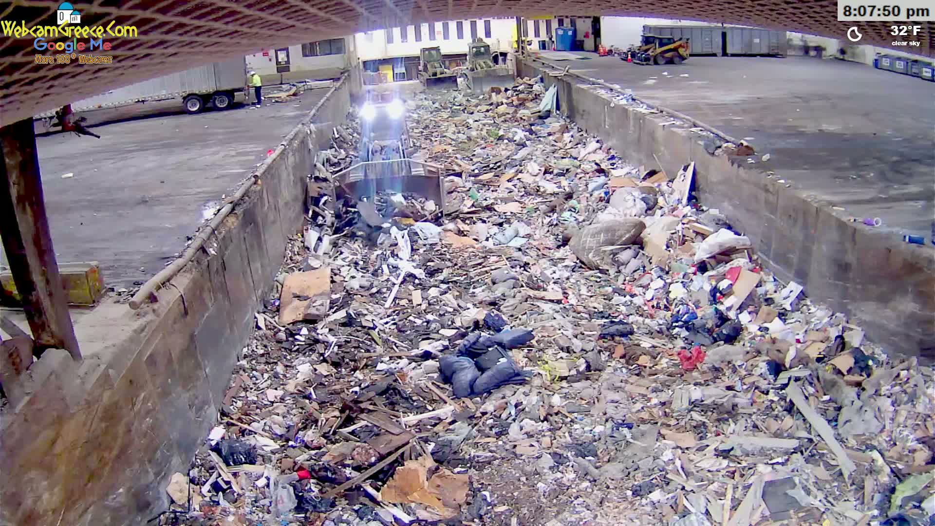A large amount of garbage is piled in a long, concrete trough under a clear sky, with machinery visible in the background.