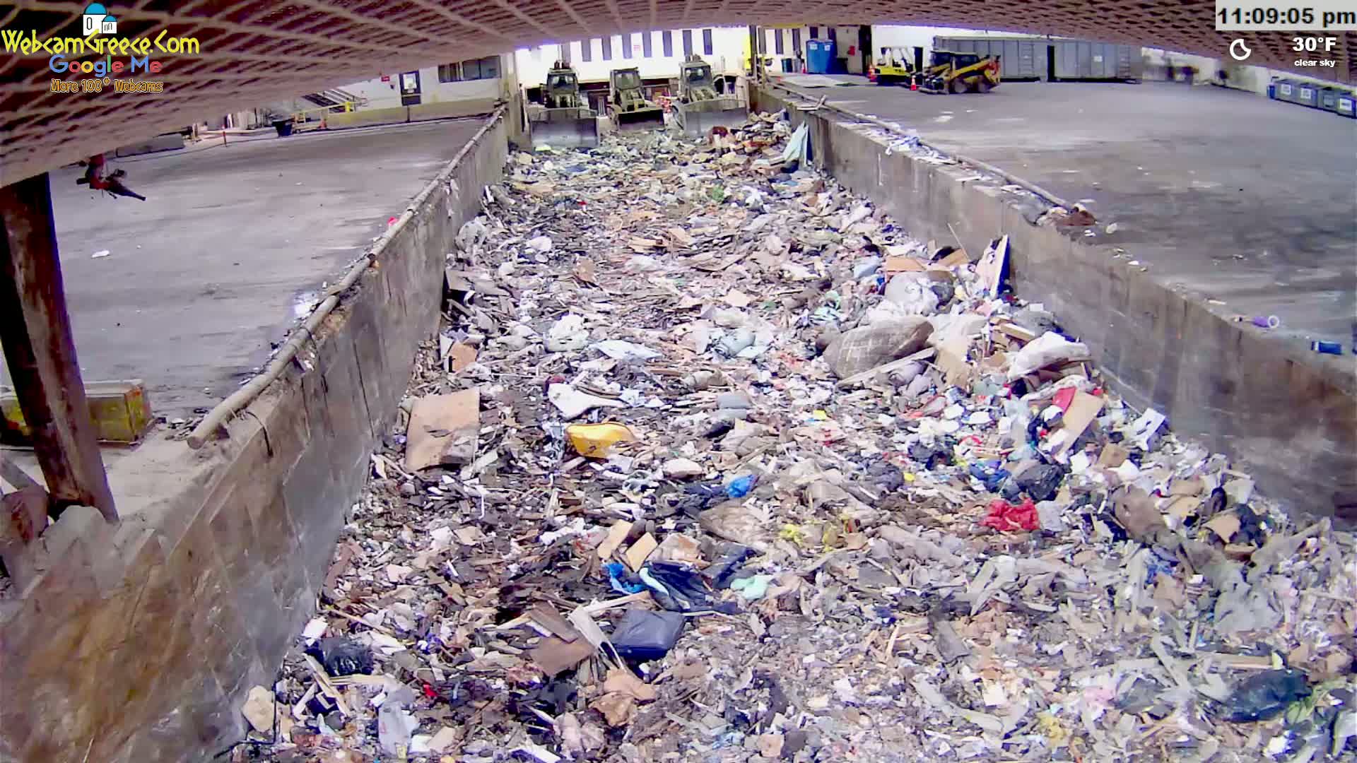A large pile of garbage sits in a concrete trough outdoors on a clear 30°F day, with two pieces of heavy machinery visible in the background.