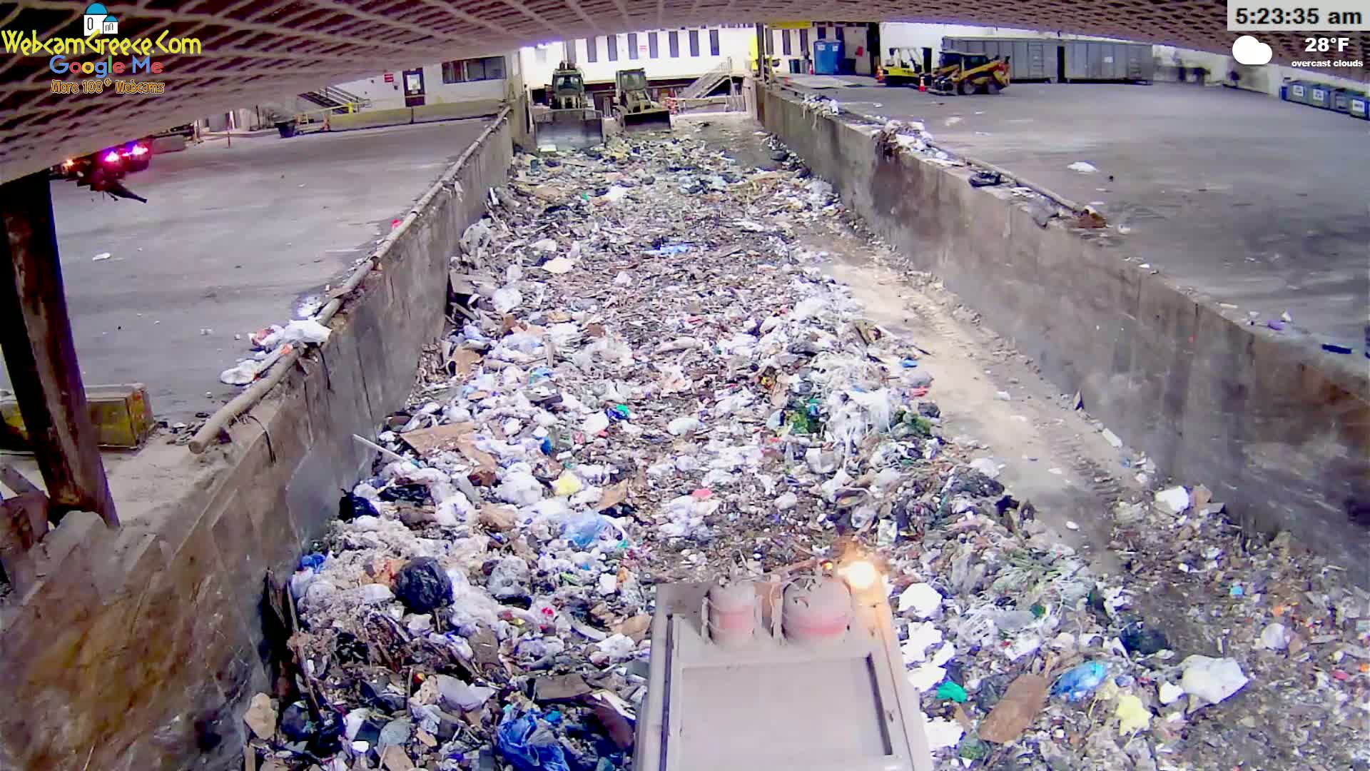 A large pile of trash fills a concrete-walled trench, with two pieces of heavy machinery visible in the background under overcast skies.