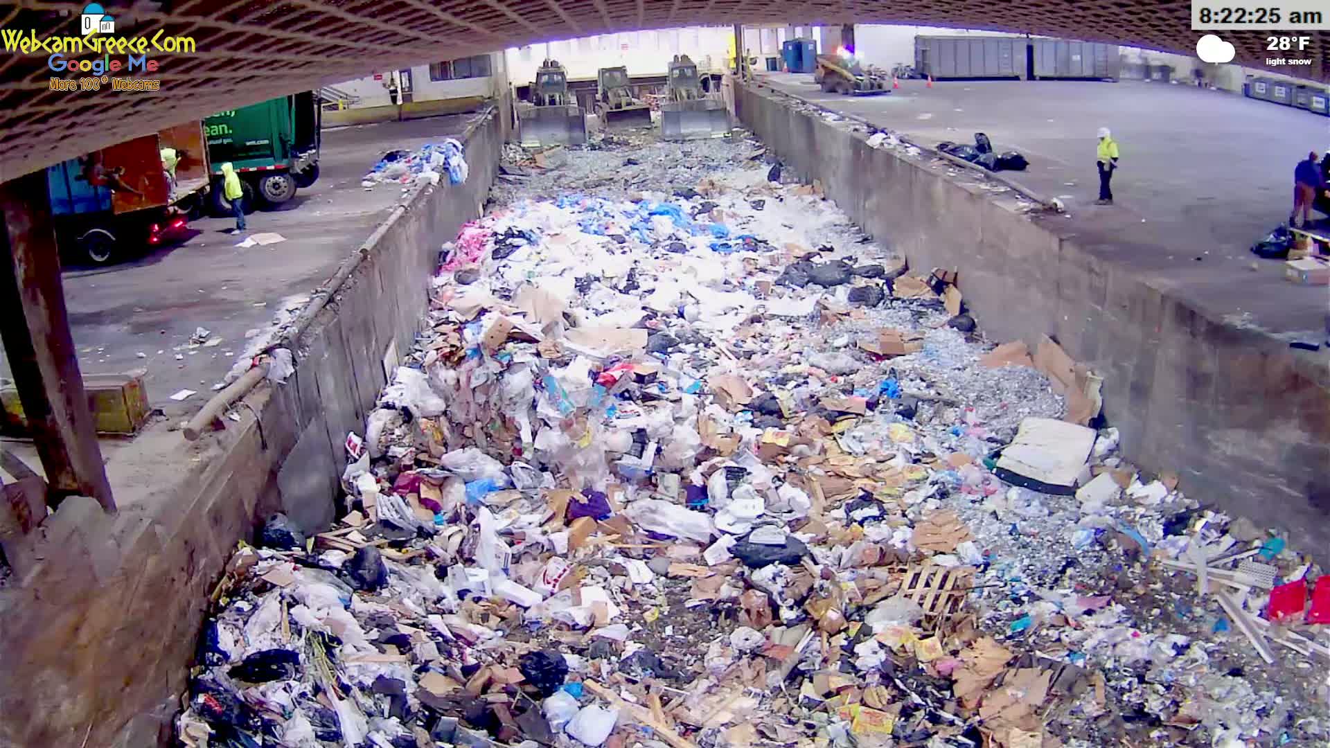 A large pile of trash and recycling is filling a concrete trough outdoors in light snow, with two loaders visible in the background and several people nearby.