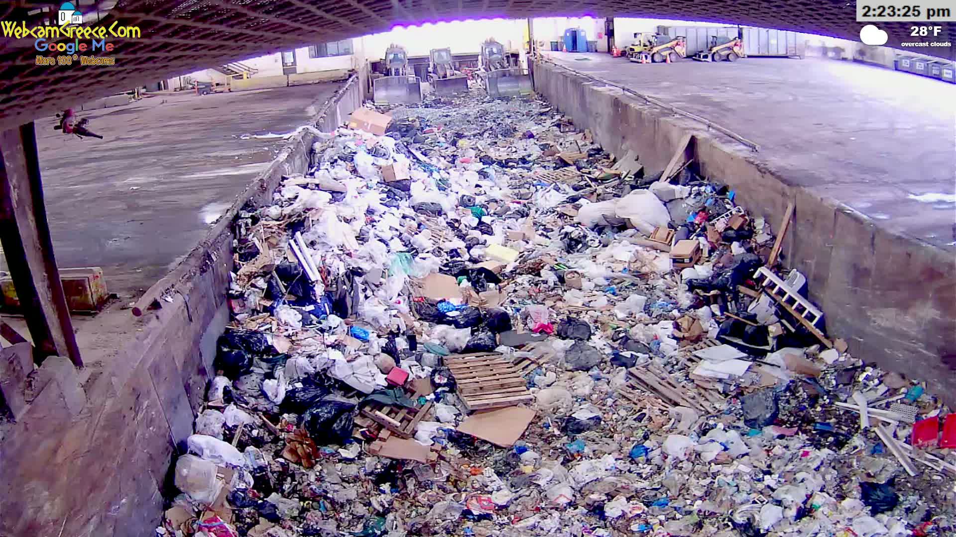 A large pile of trash sits in a concrete-walled receptacle under overcast skies, with three pieces of heavy machinery visible in the background.