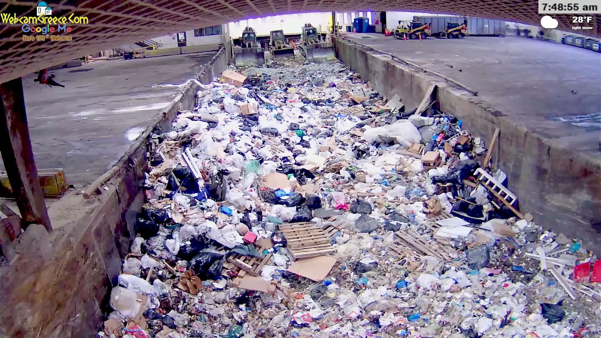 A large pile of garbage sits in a concrete trough under a partly cloudy sky, with two pieces of heavy machinery visible in the background.