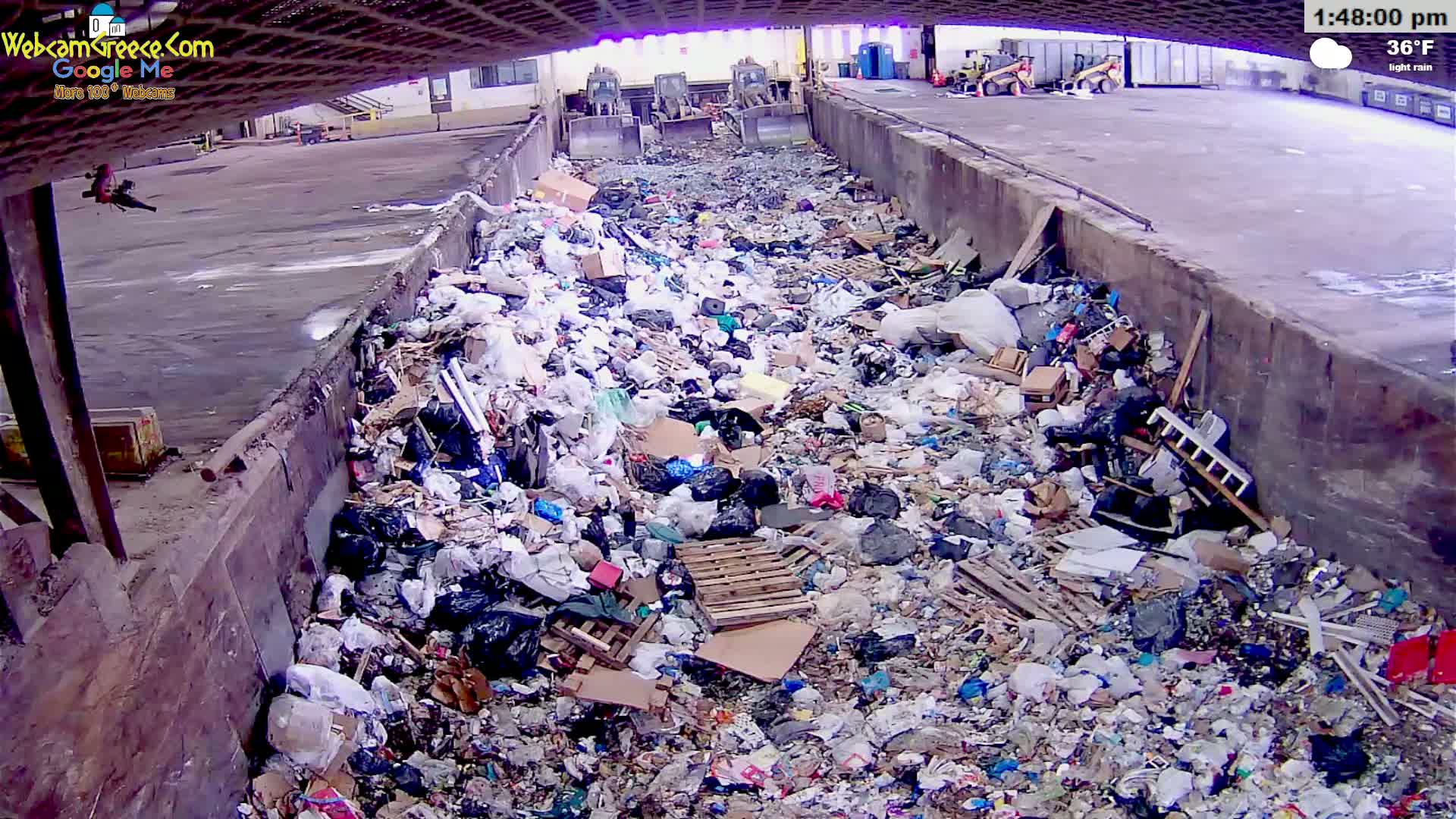 A large pile of garbage sits in a concrete trough outdoors in light rain, with some construction equipment visible in the background.