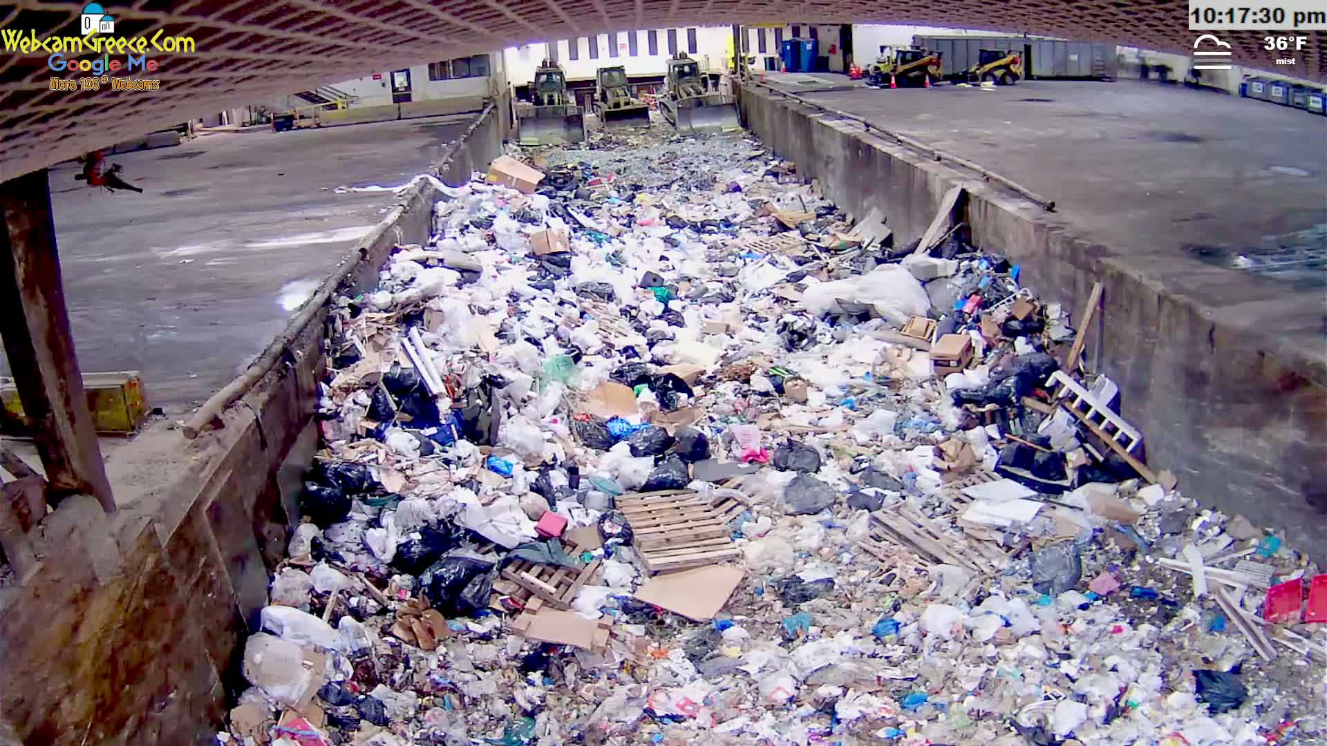 A large pile of garbage sits in a concrete trough outdoors in misty 36°F weather, with construction equipment visible in the background.