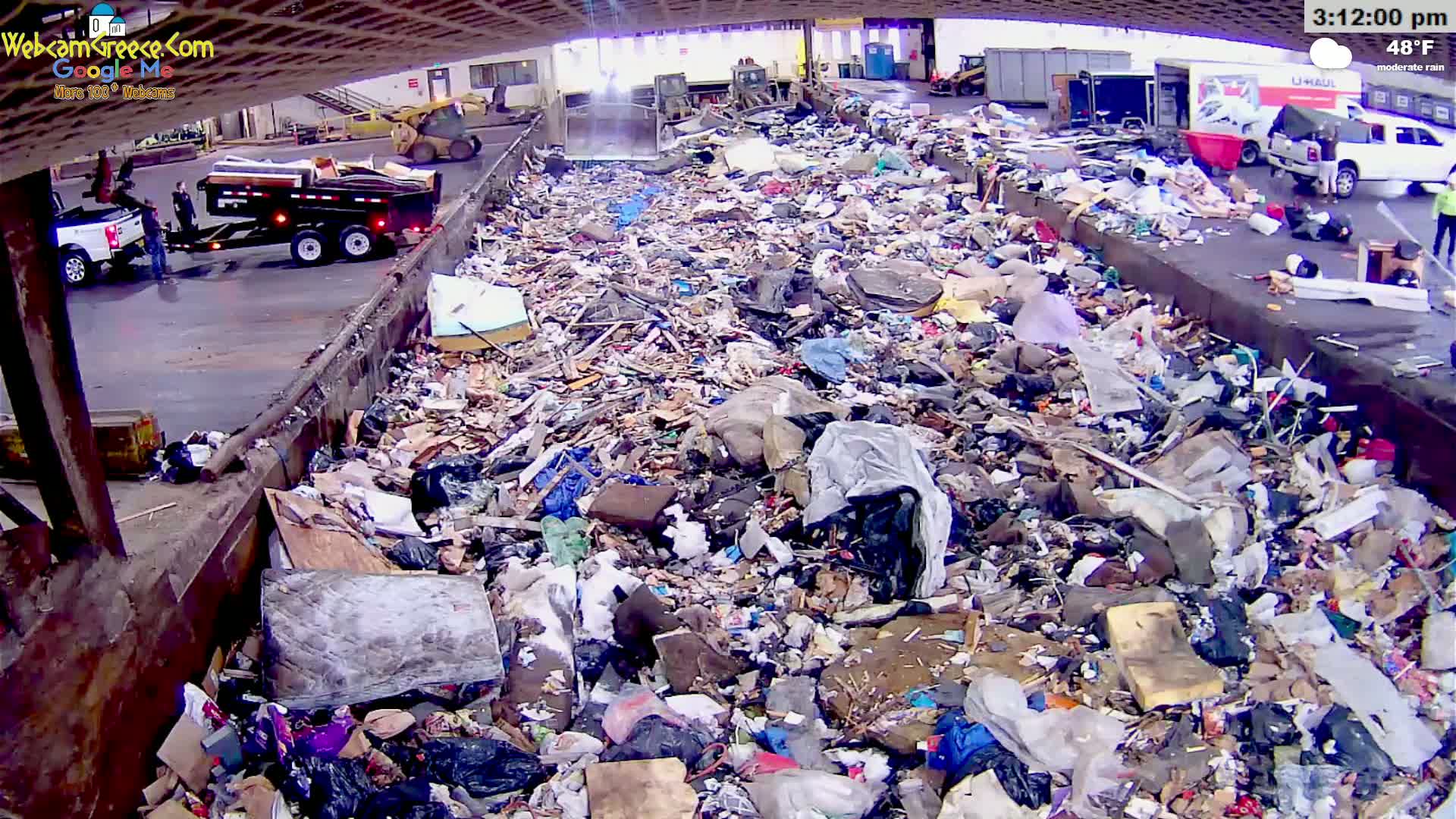 A large pile of refuse sits inside a dumpster in moderate rain.