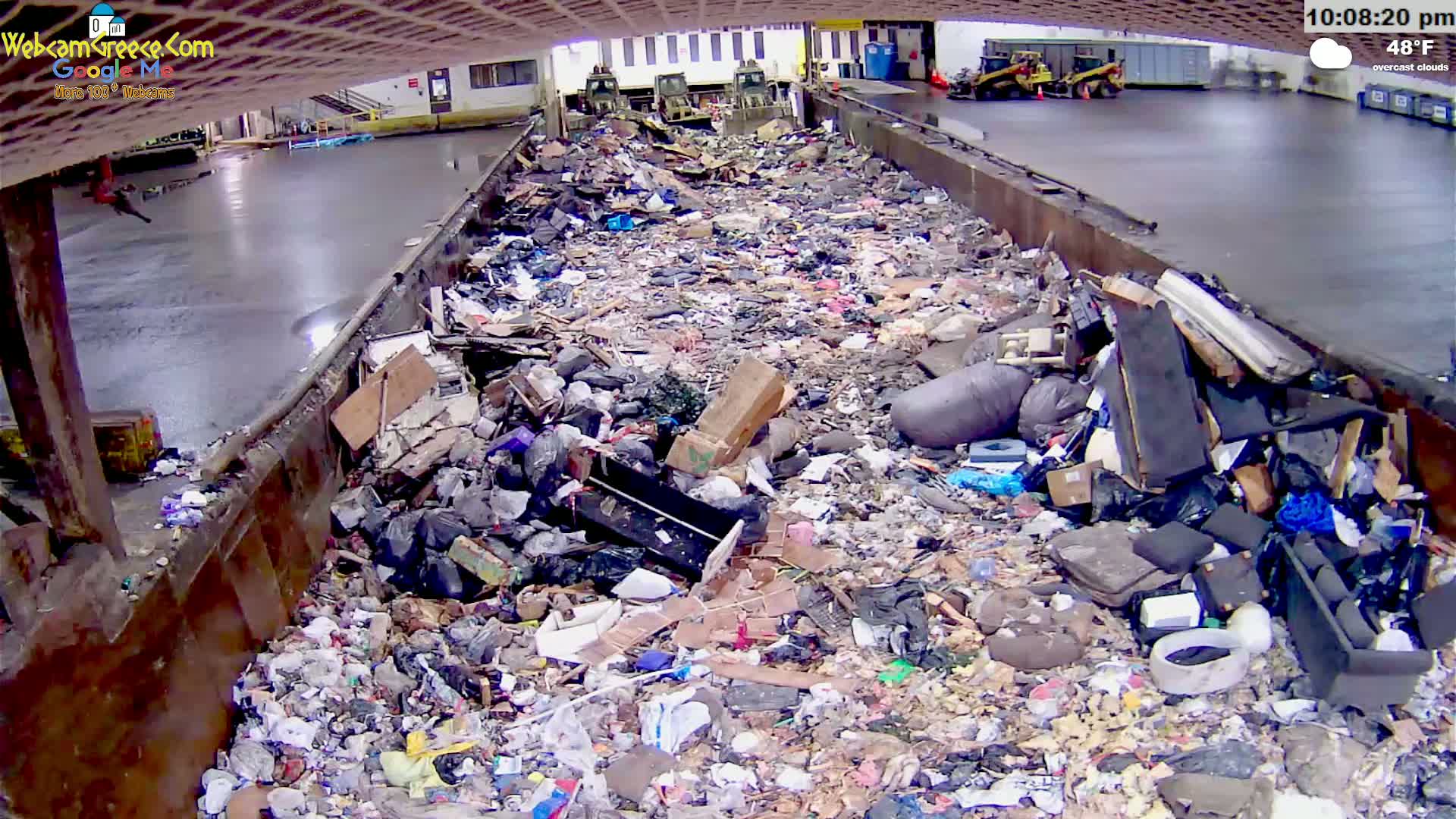 A large, overflowing dumpster filled with various types of trash is shown in an indoor facility under overcast skies.
