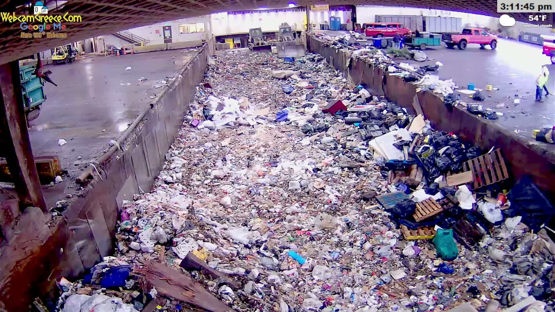 A large amount of trash and debris fills a wide, rusty trough outdoors under overcast skies.