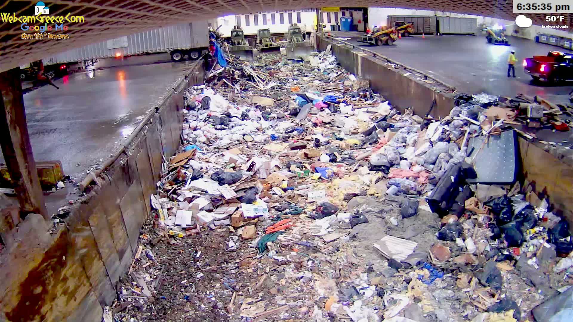 A large pile of garbage is seen filling a concrete trough under an overcast sky, with trucks and machinery visible in the background.