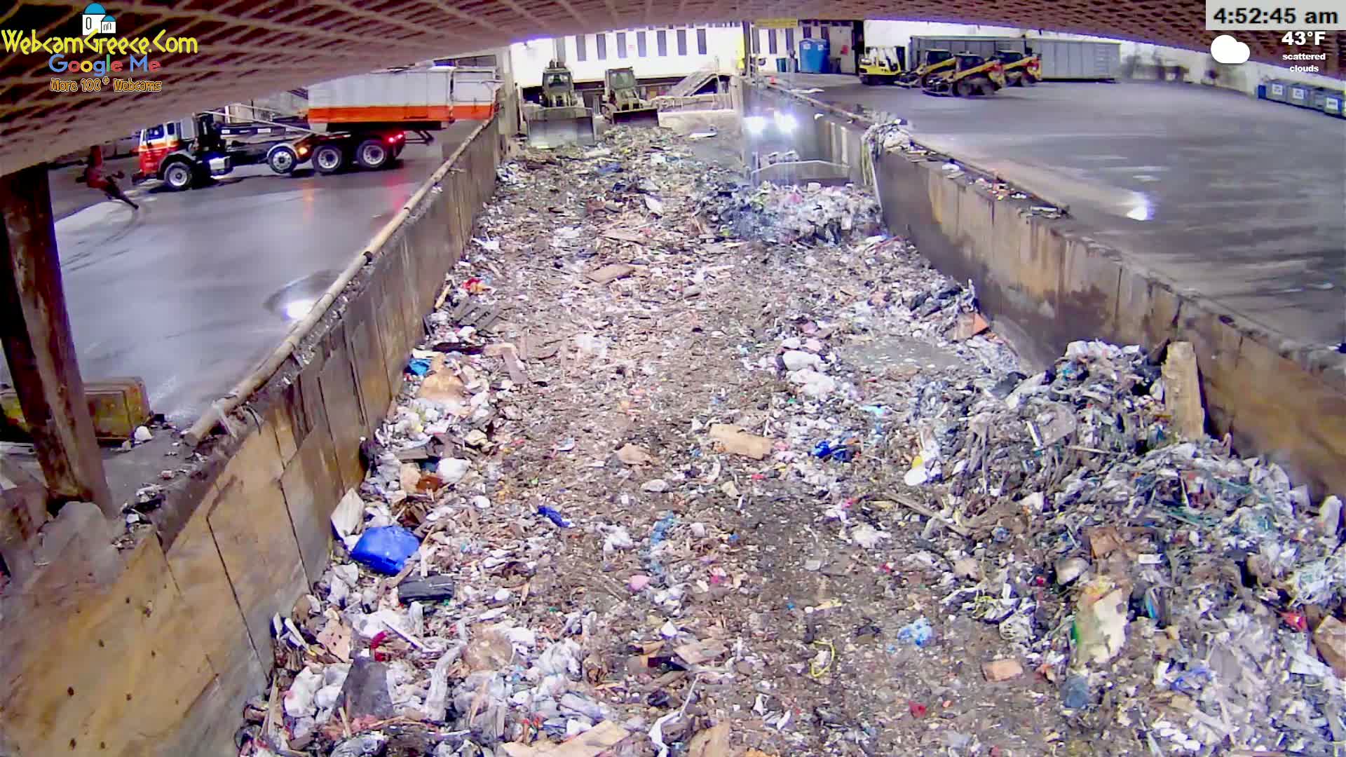 A large pile of trash fills a long, narrow concrete trough under overcast skies, with two garbage trucks and construction equipment visible in the background.