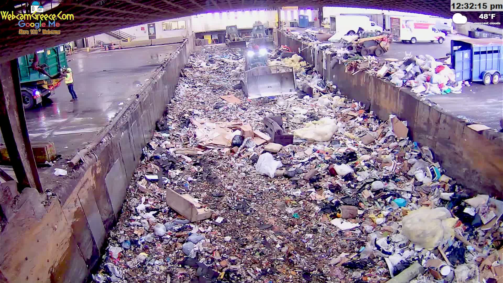A large amount of garbage is piled in a concrete trough under overcast skies, with a small bulldozer working inside the pile and a person standing near a garbage truck outside.