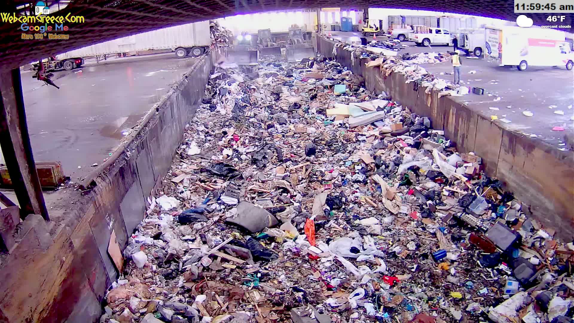 A large pile of trash is seen filling a concrete-walled pit on an overcast day, with trucks and workers visible in the background.