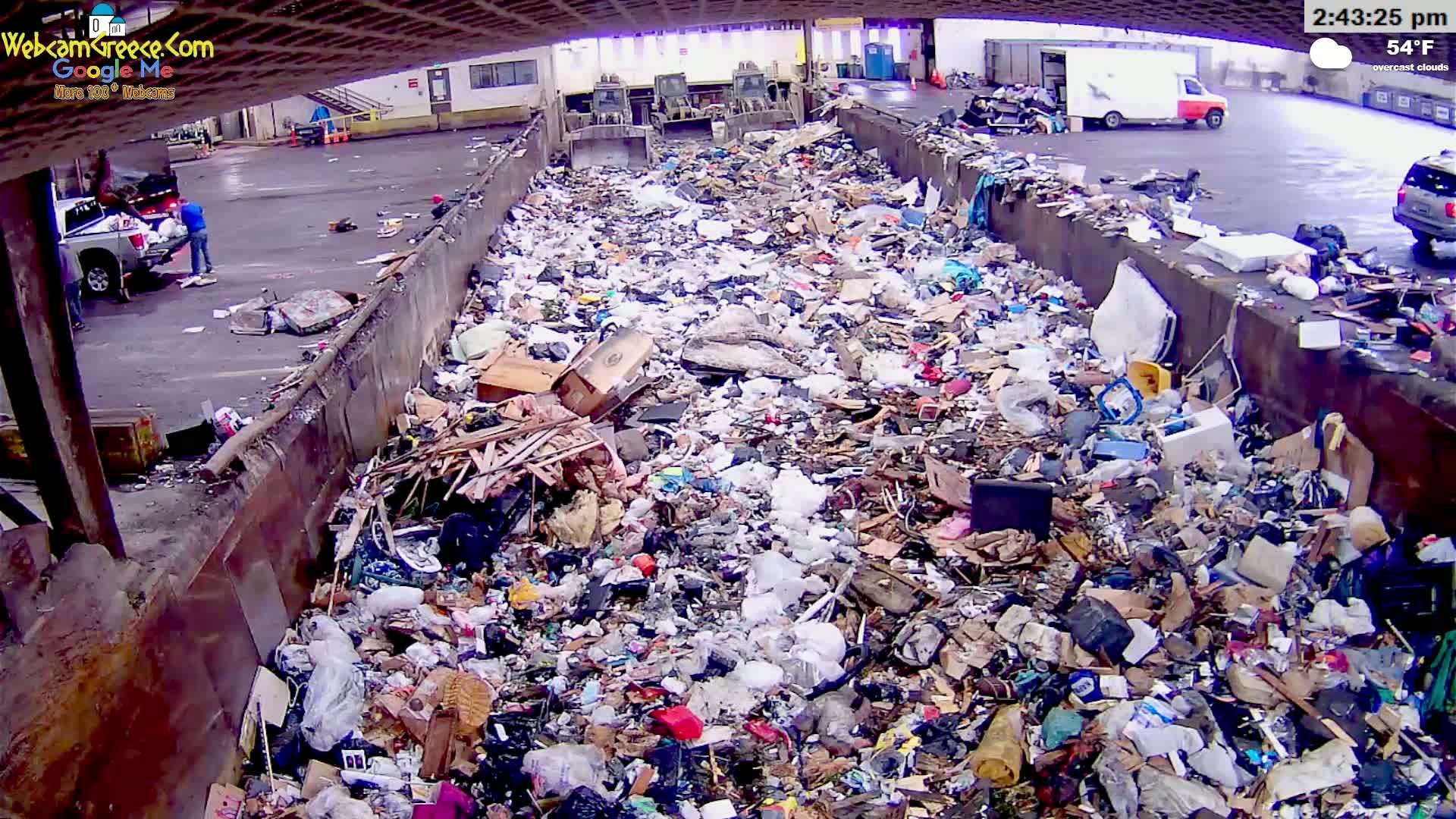 A large dumpster is overflowing with a diverse assortment of trash under overcast skies.