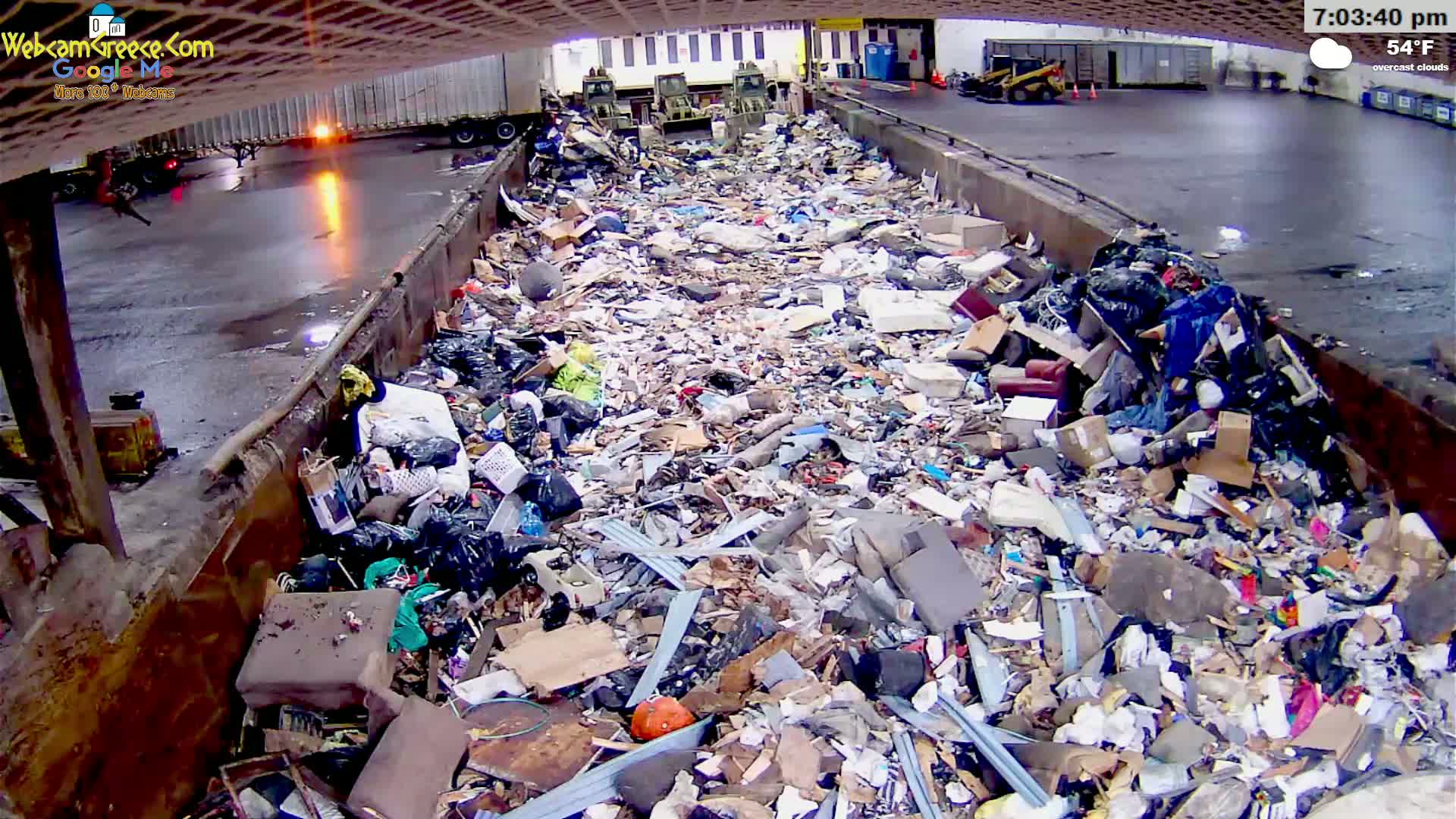 A large dumpster is overflowing with a diverse assortment of garbage under overcast skies at 54°F.