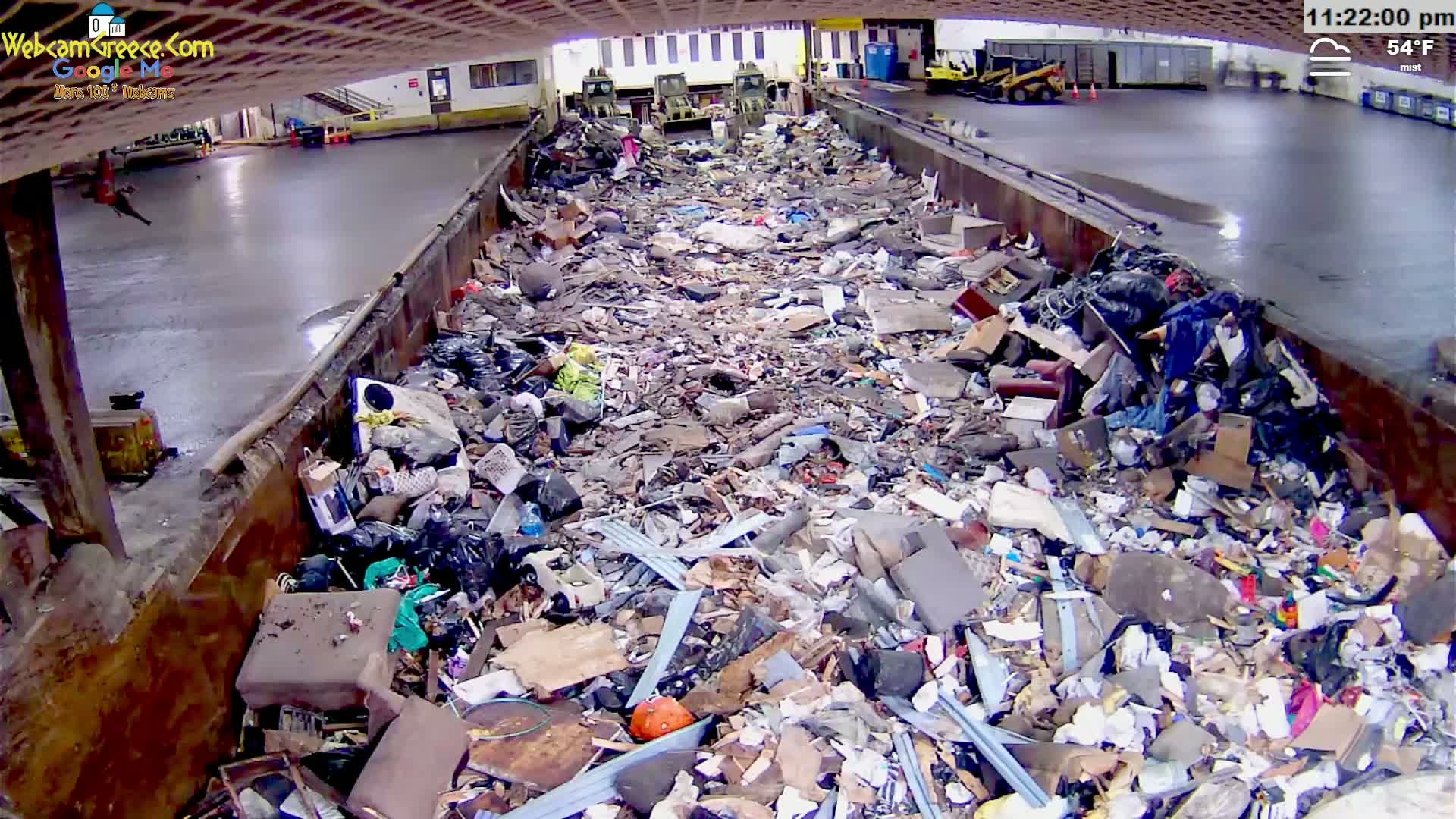A large metal trough is filled with a massive pile of mixed garbage under misty, 54°F conditions.