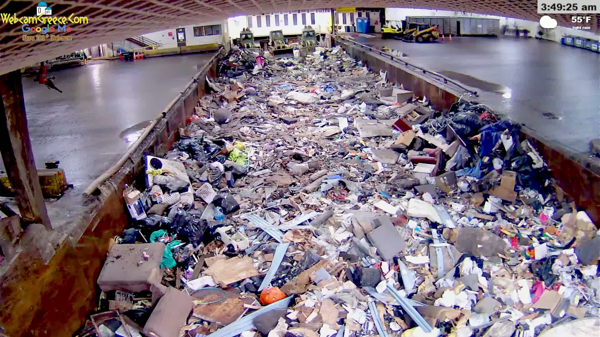 A large metal container is filled to the brim with a massive pile of assorted trash and debris under a lightly raining sky.