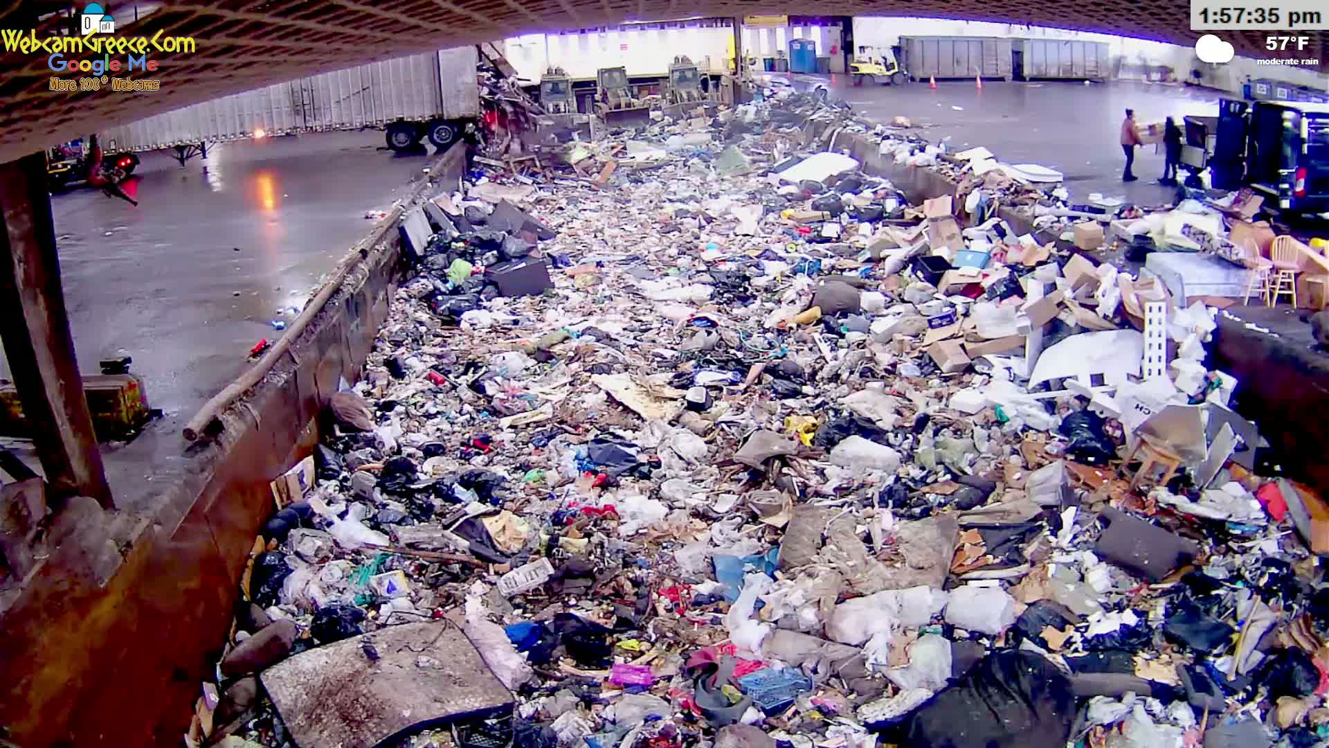 A large pile of refuse is seen under an overpass in moderate rain.